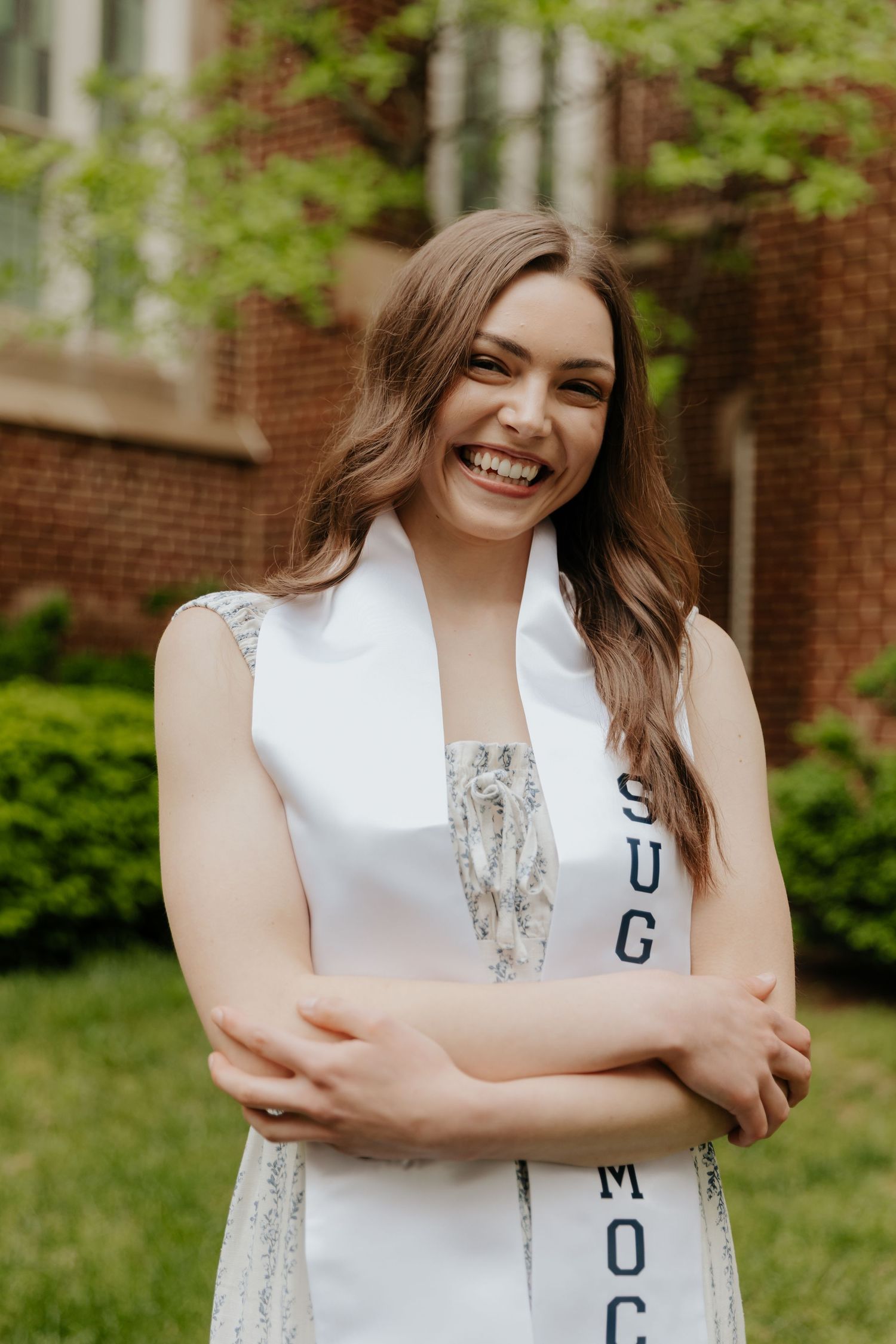 Graduate in white stole with silver lettering poses outdoors near brick buildings.