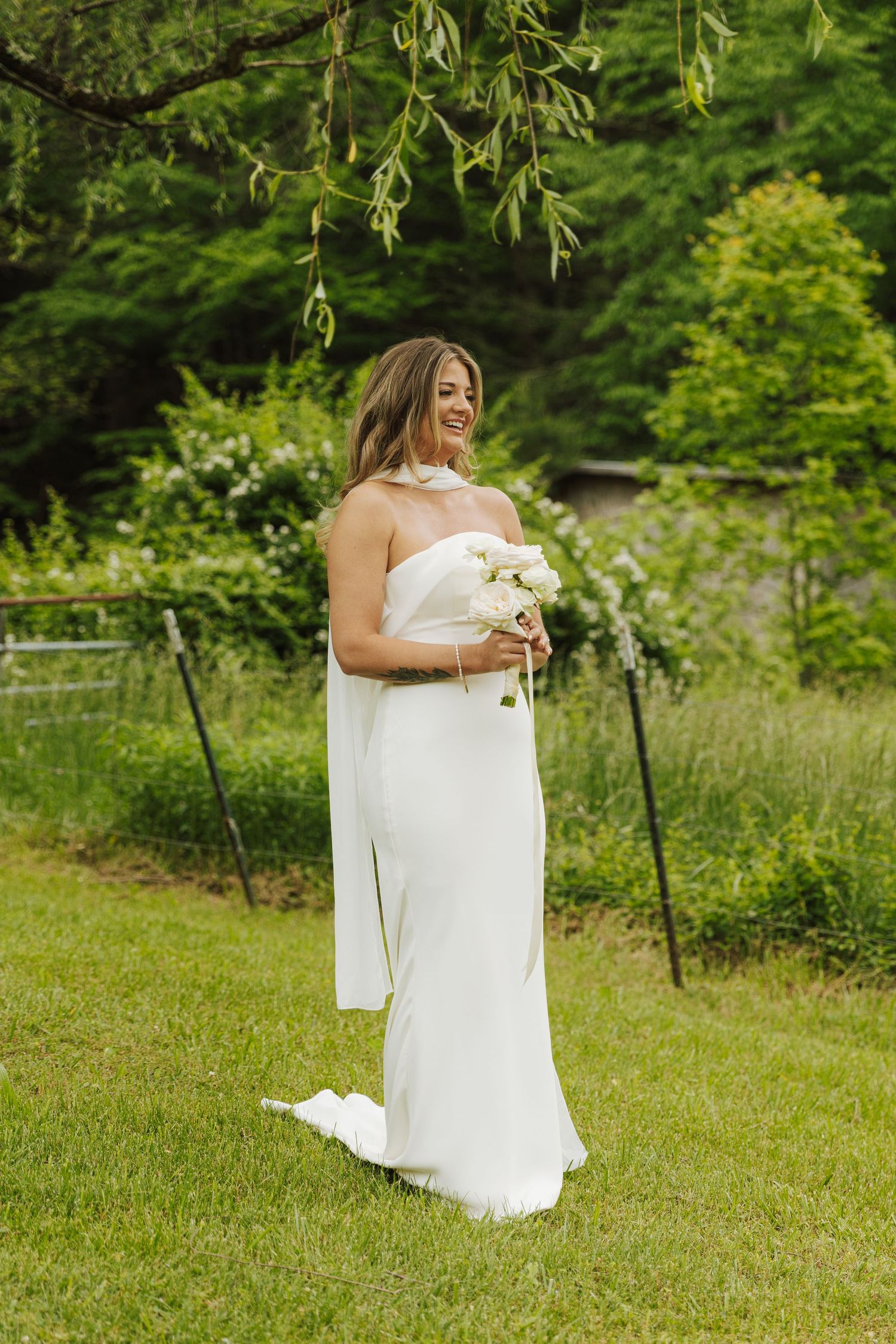 Person in white dress holding bouquet stands in lush garden setting.