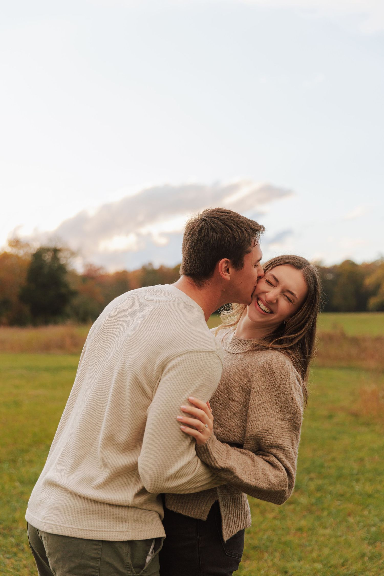 A couple laughs and embraces while standing in a scenic meadow on a bright autumn day.