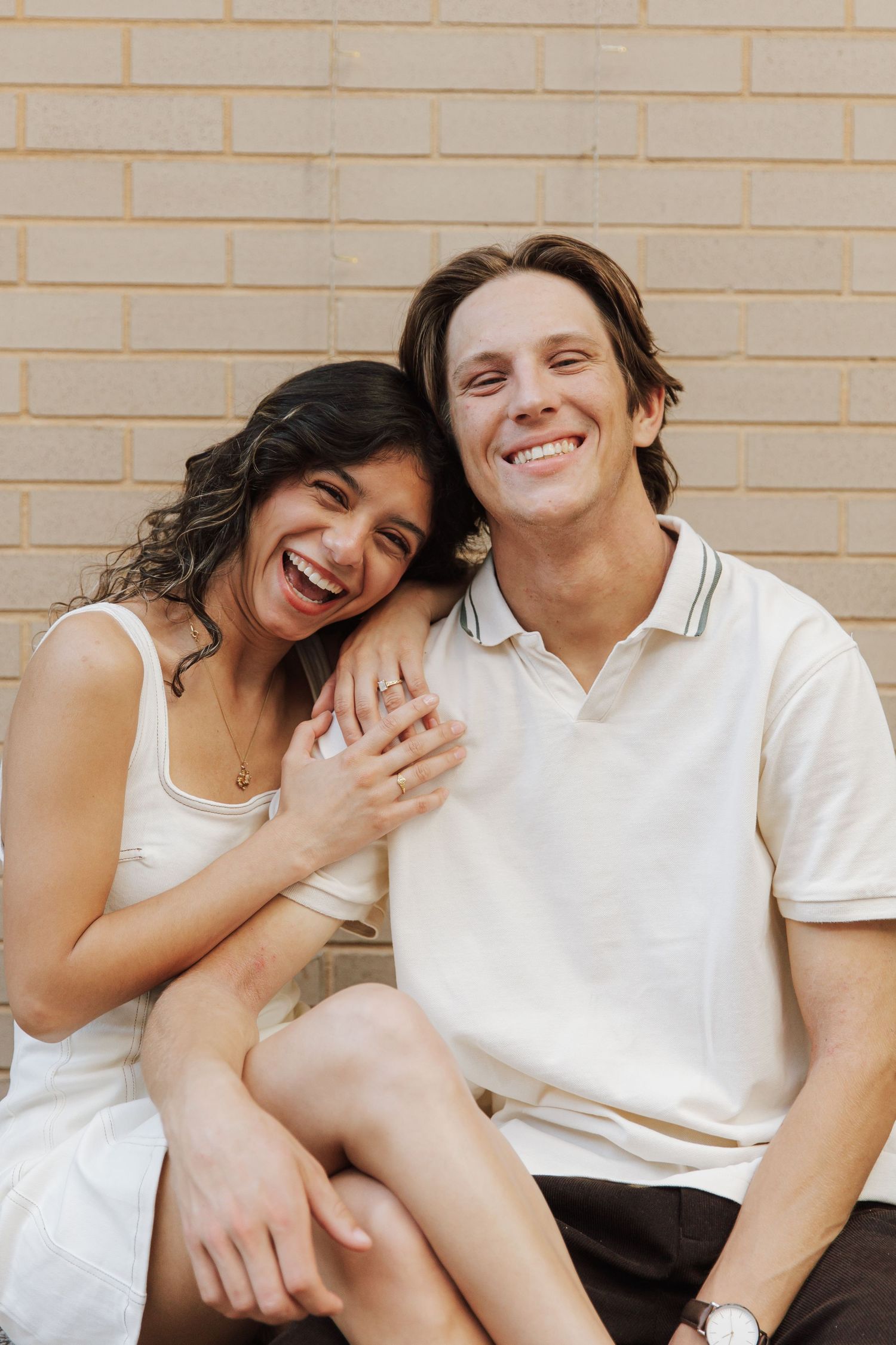 A joyful couple laughing together against a brick wall, woman in white dress leaning on mans shoulder.