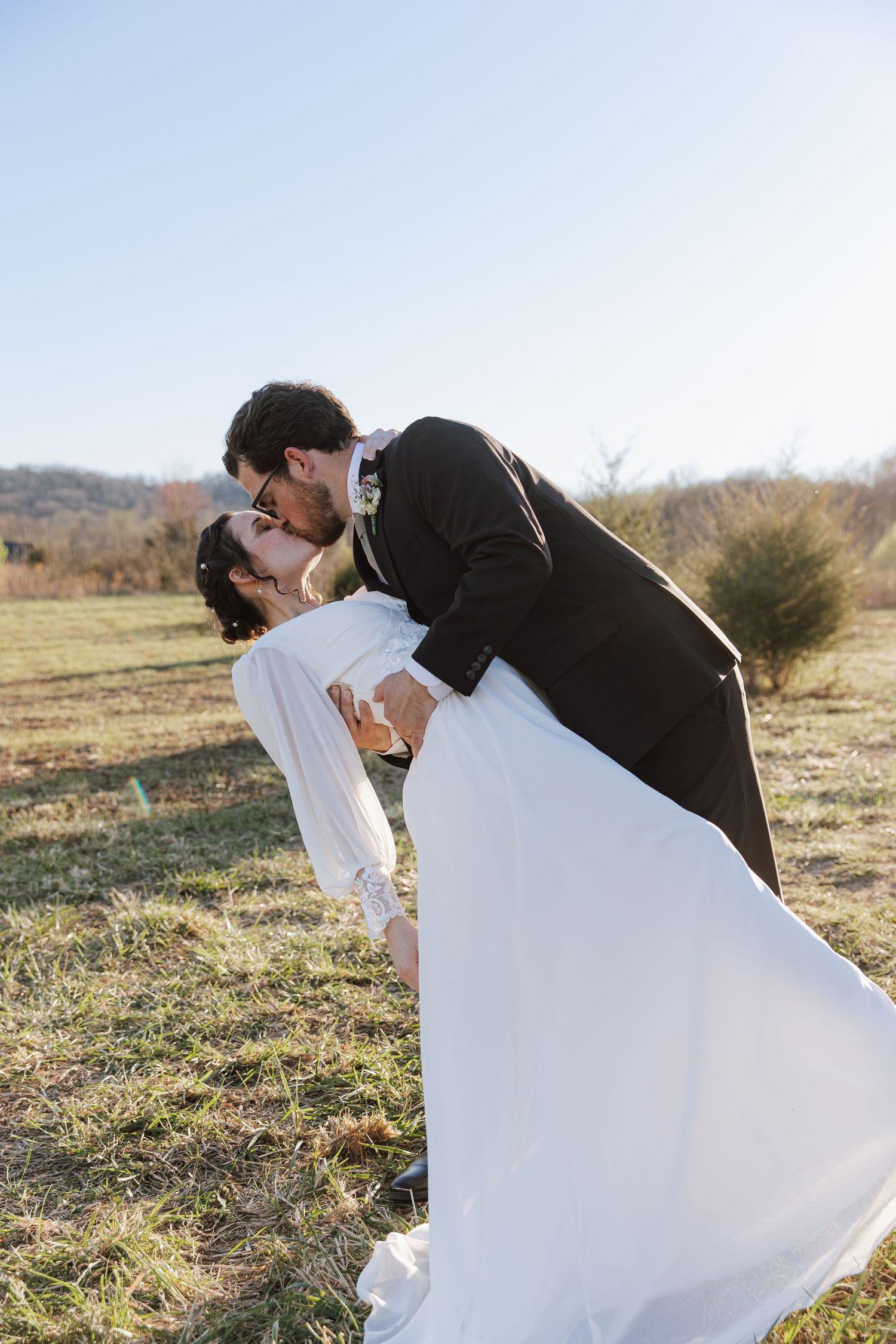 Groom dipping and kissing bride in flowing white dress in sunny open field on their wedding day.