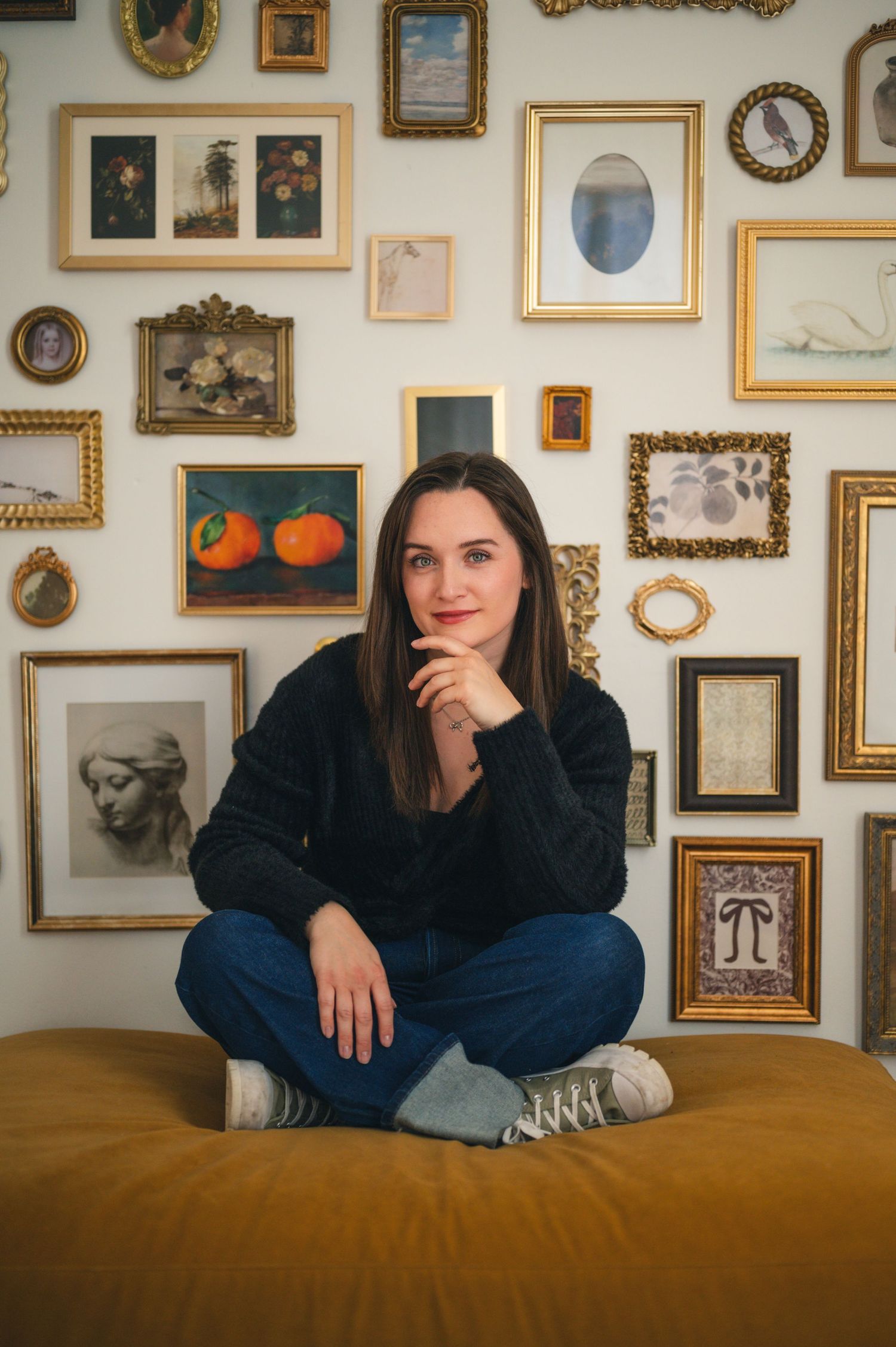 Brunette woman sitting cross-legged on ottoman in front of eclectic gallery wall with ornate frames.