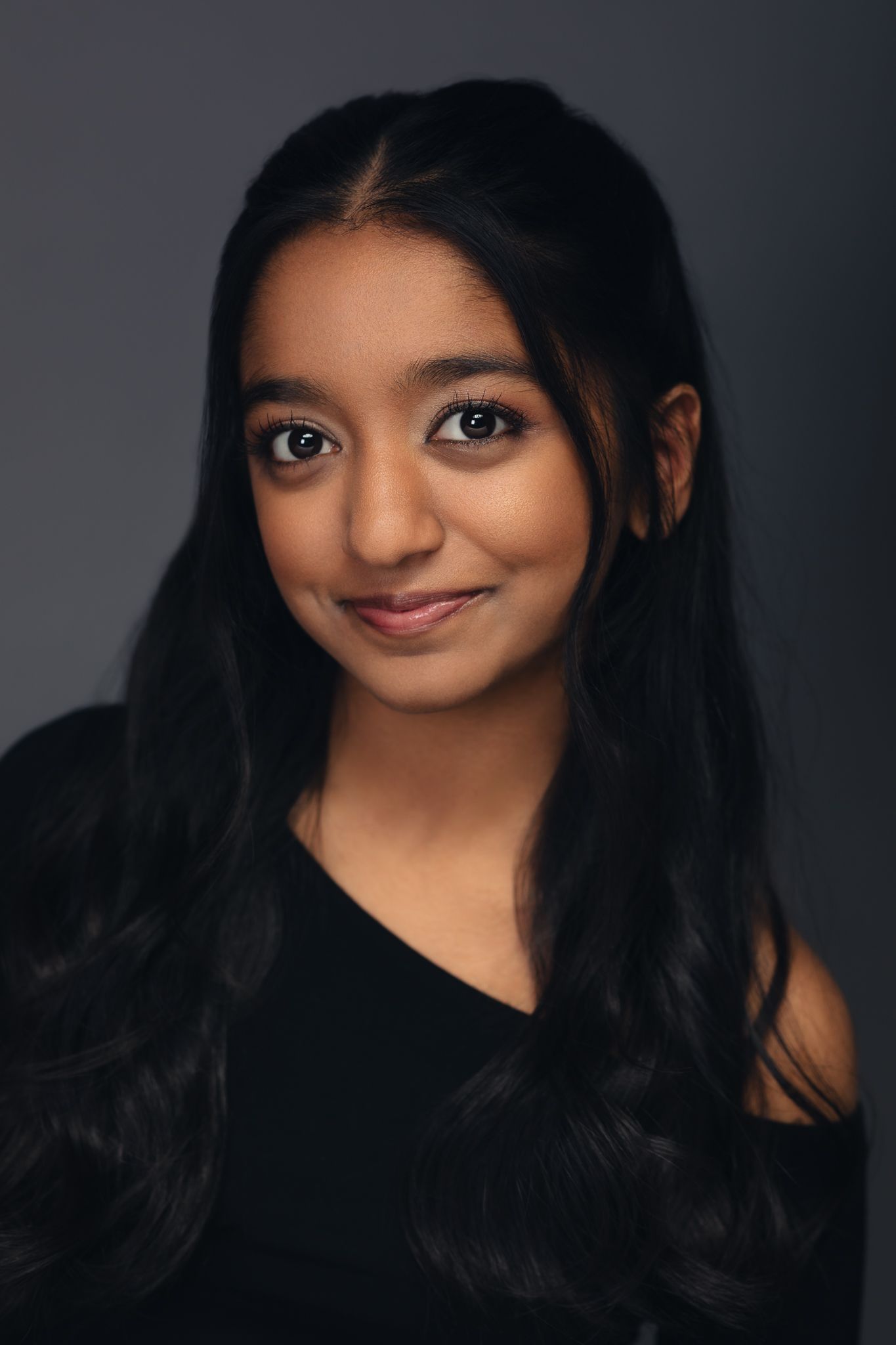 Young woman with long black hair smiling softly in black top against dark gray background.