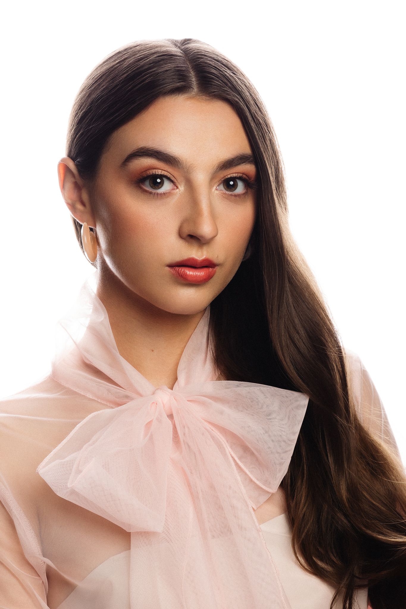 Young woman with long dark hair wearing a pink bow blouse and gold hoop earring against white background.