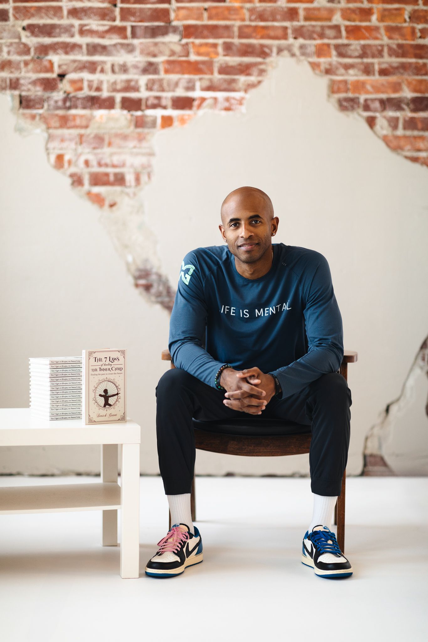Man in blue 'Life is Mental' sweatshirt sits on chair near books against rustic brick wall.
