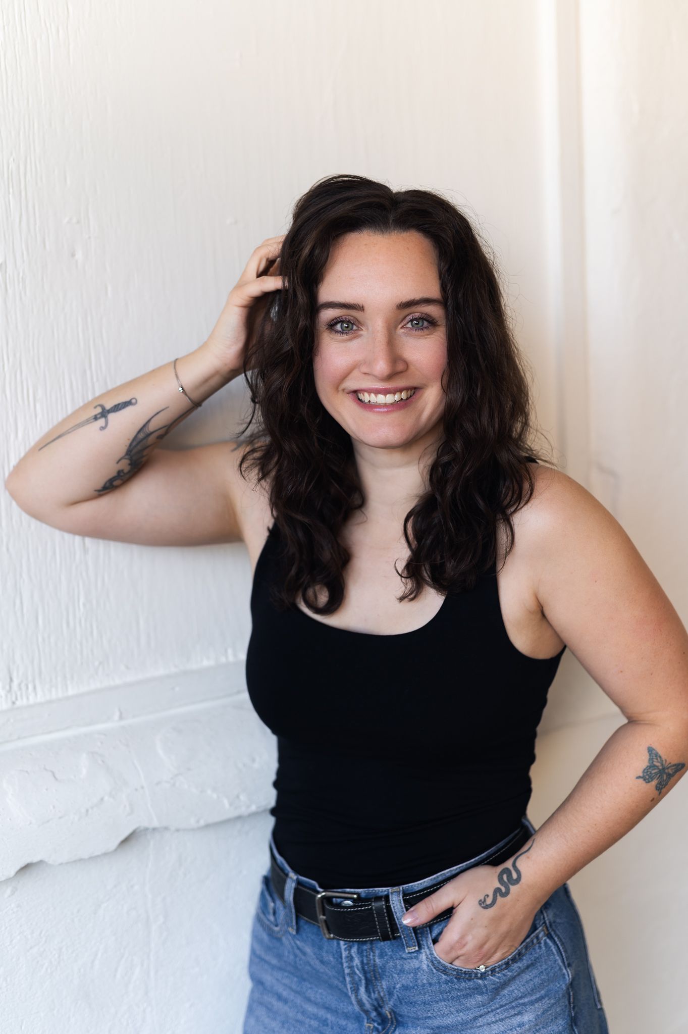 Smiling woman with dark curly hair, tattoos, black tank top, and jeans posing against a white wall.