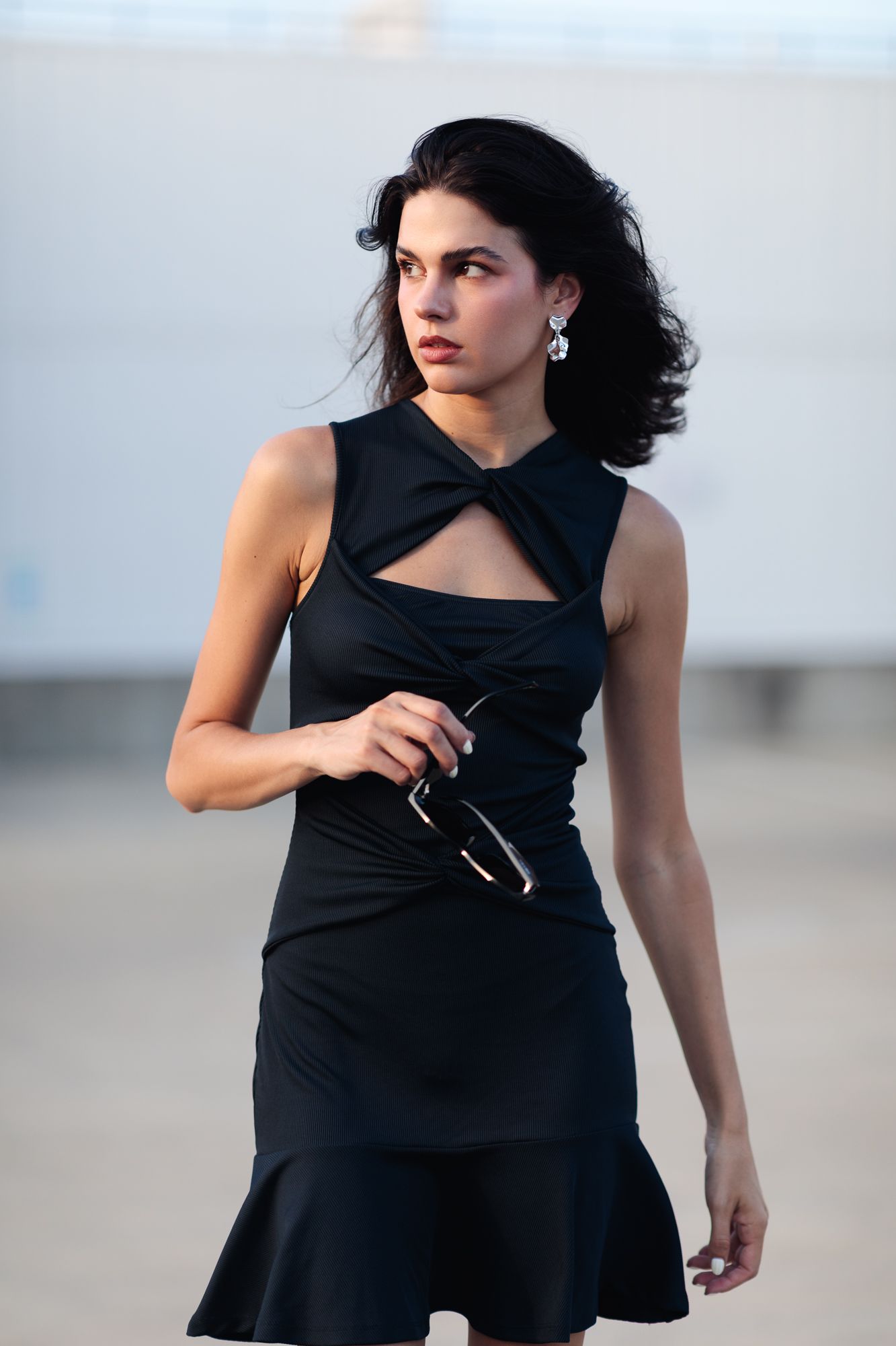 Woman in black cutout mini dress holding sunglasses outdoors.