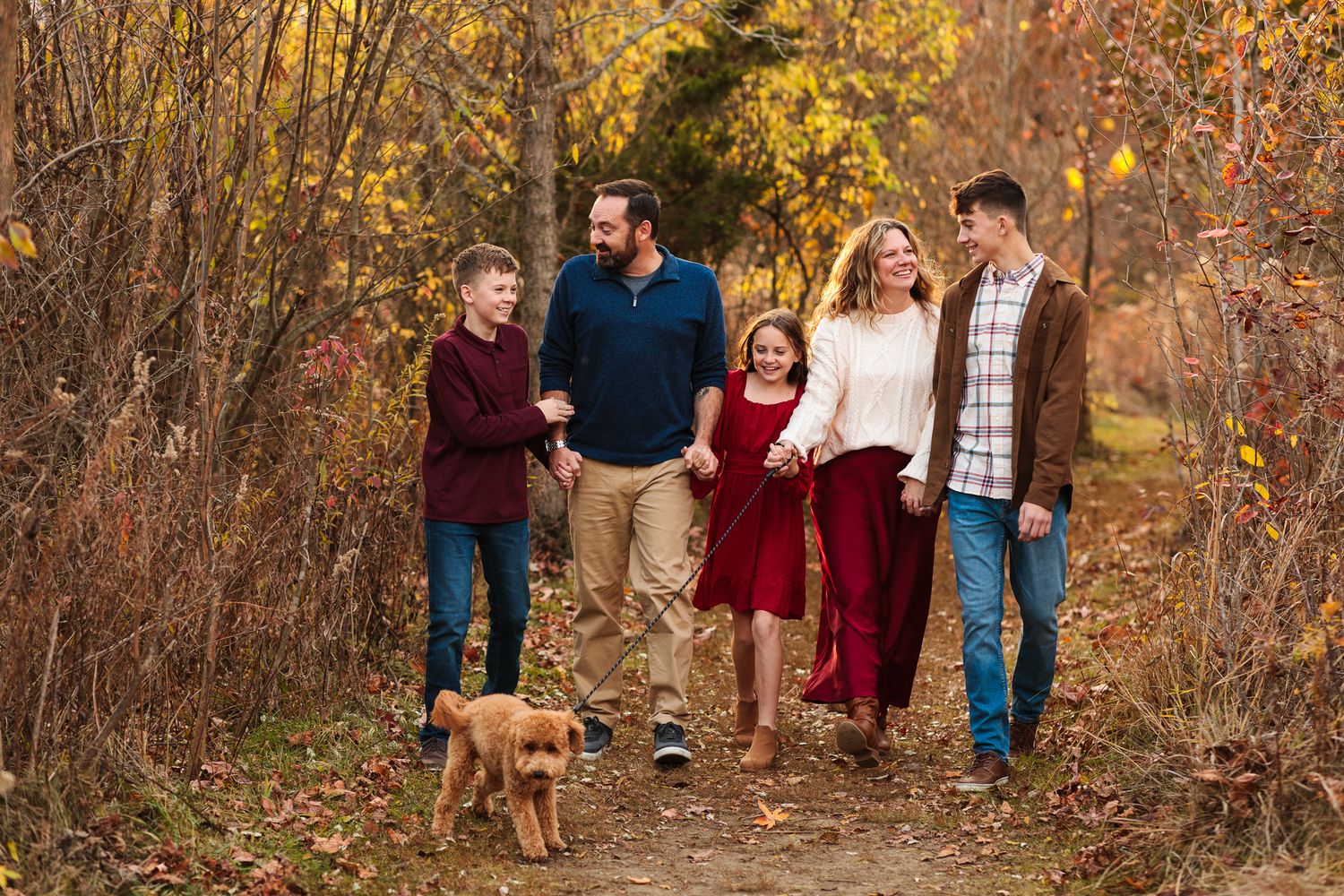 Happy family of five walking dog on autumn forest path surrounded by colorful fall foliage.
