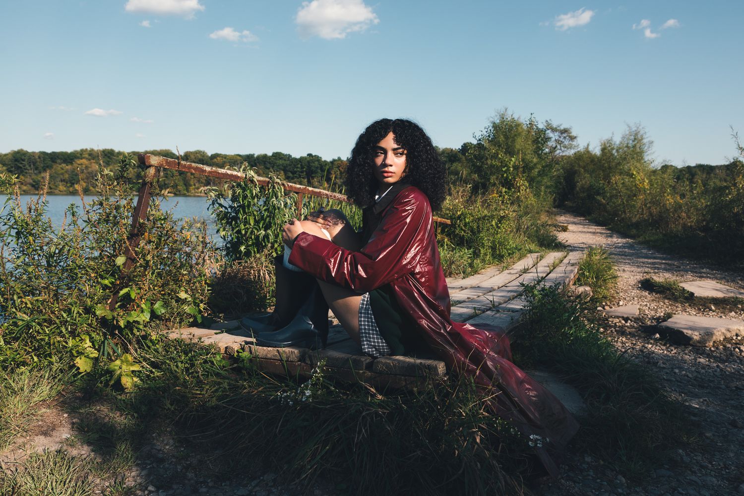 Young woman in burgundy jacket sitting on logs near a lake in natural outdoor setting.