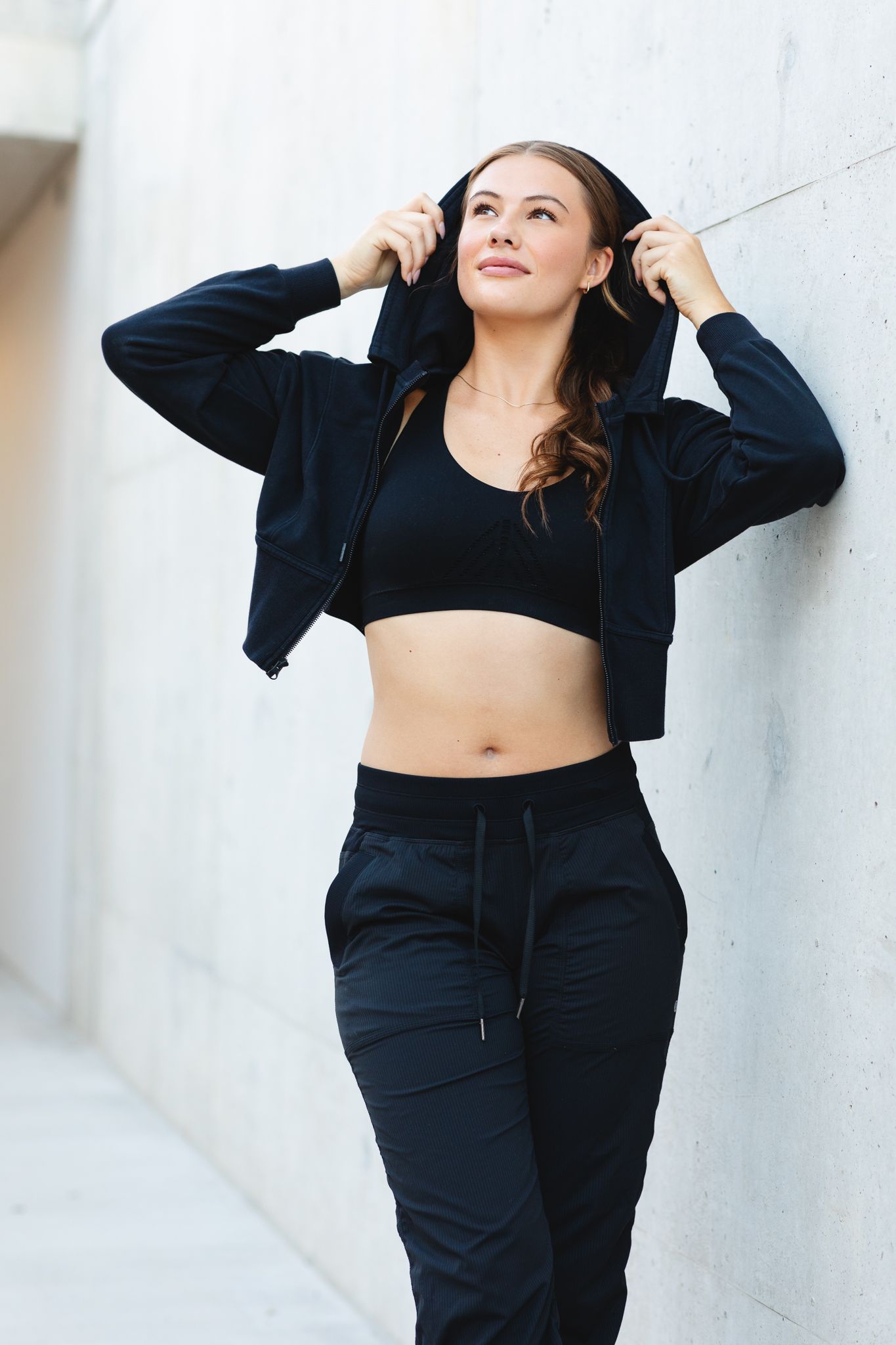 Fit woman in black sports bra and hoodie leaning against concrete wall outdoors.