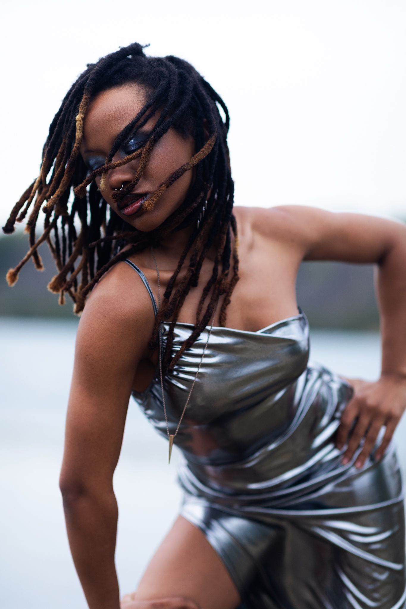 Woman with dreadlocks wearing a silver metallic strapless dress poses outdoors near water.