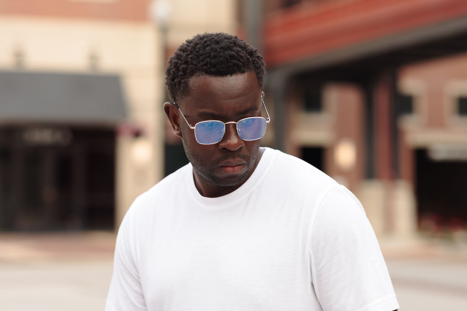 Man in white t-shirt and blue mirrored sunglasses standing outdoors on urban street.