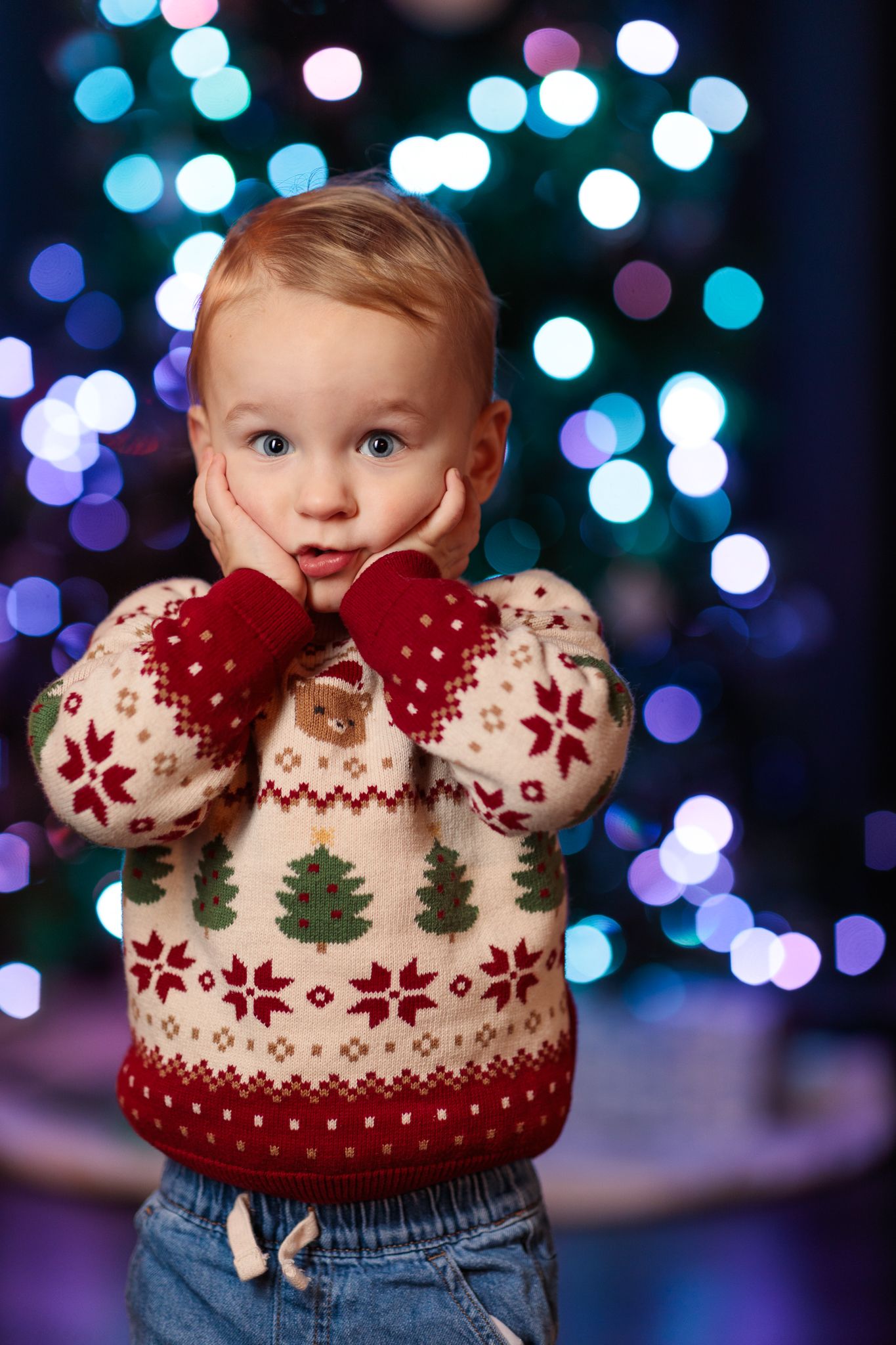 Surprised toddler in a Christmas sweater and mittens holding cheeks with wide eyes against bokeh holiday lights background.