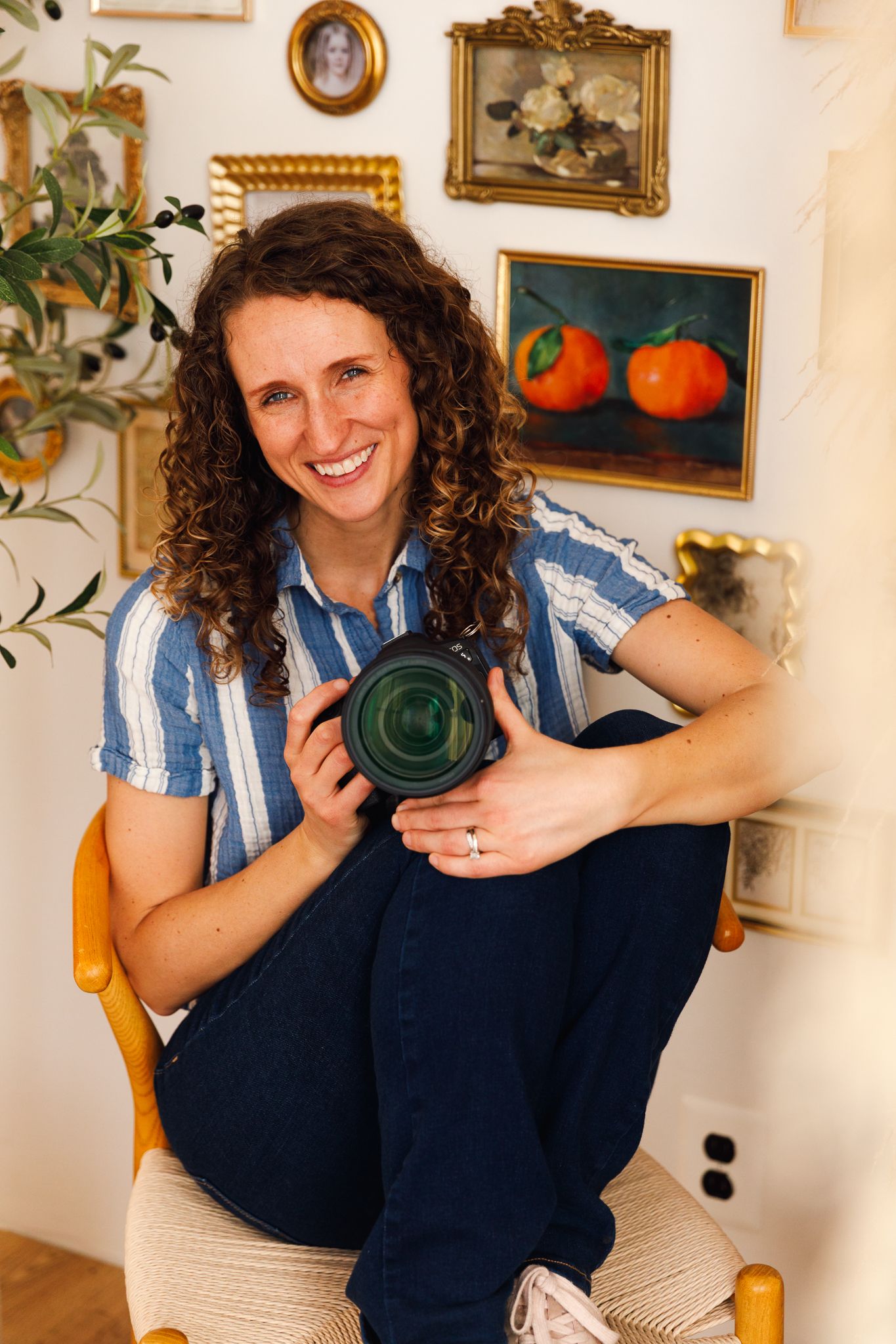 Smiling woman with curly hair holding a camera lens, sitting in a chair surrounded by vintage framed artwork on a white wall.