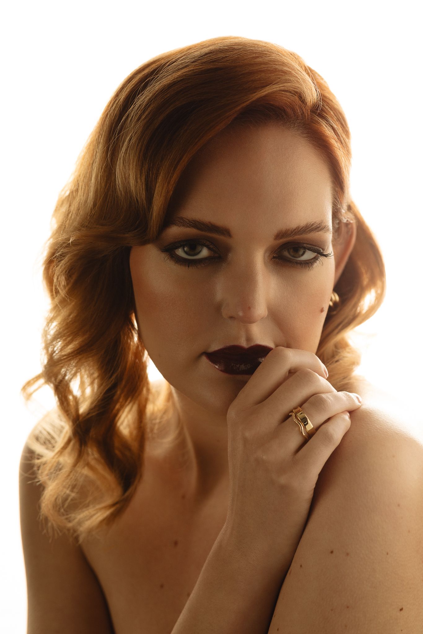 Redhead woman with dark lips and smoky eyes posing with hand near face against white background.
