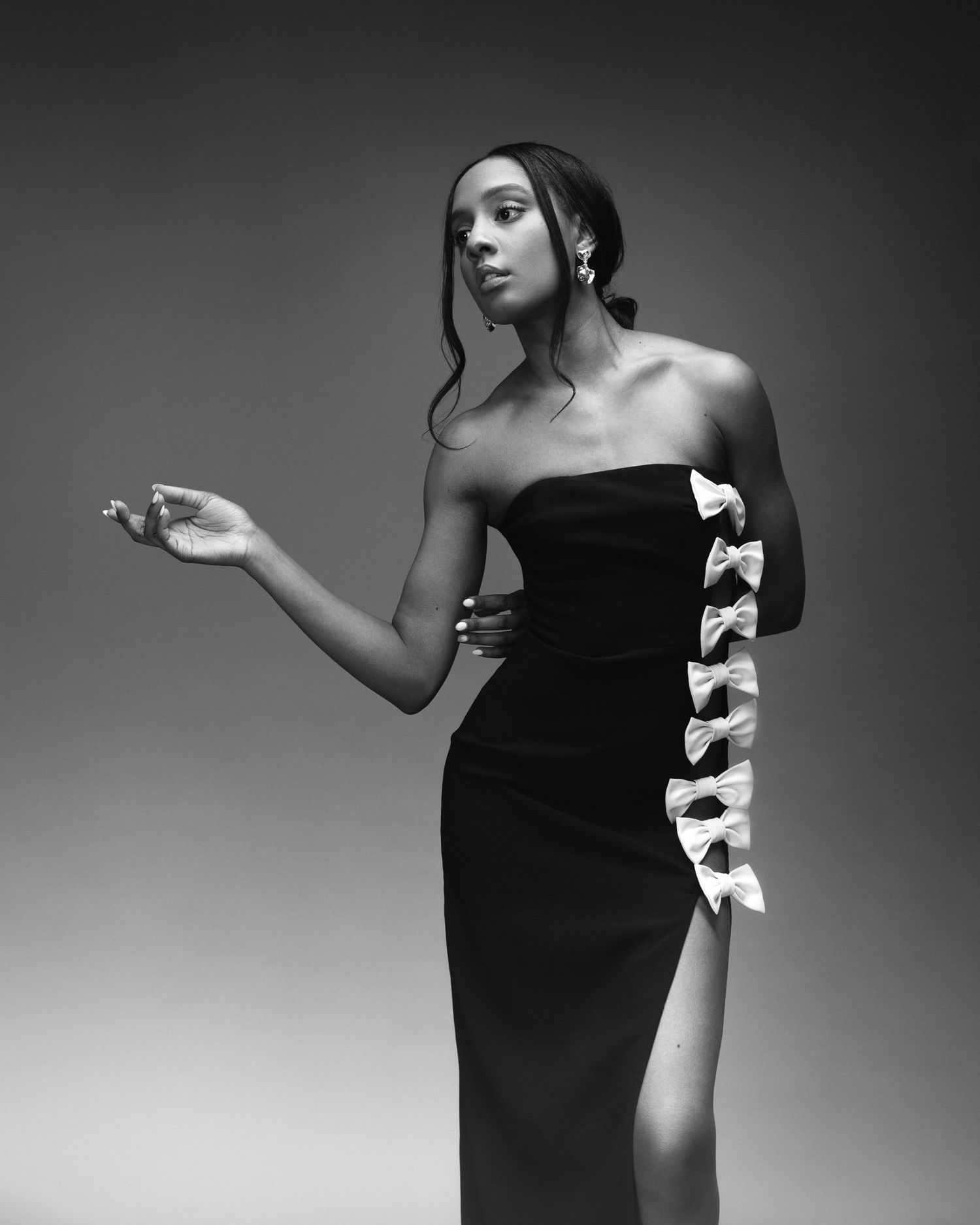 Black and white photo of a woman in a strapless gown with bow details and a slit, posing elegantly.