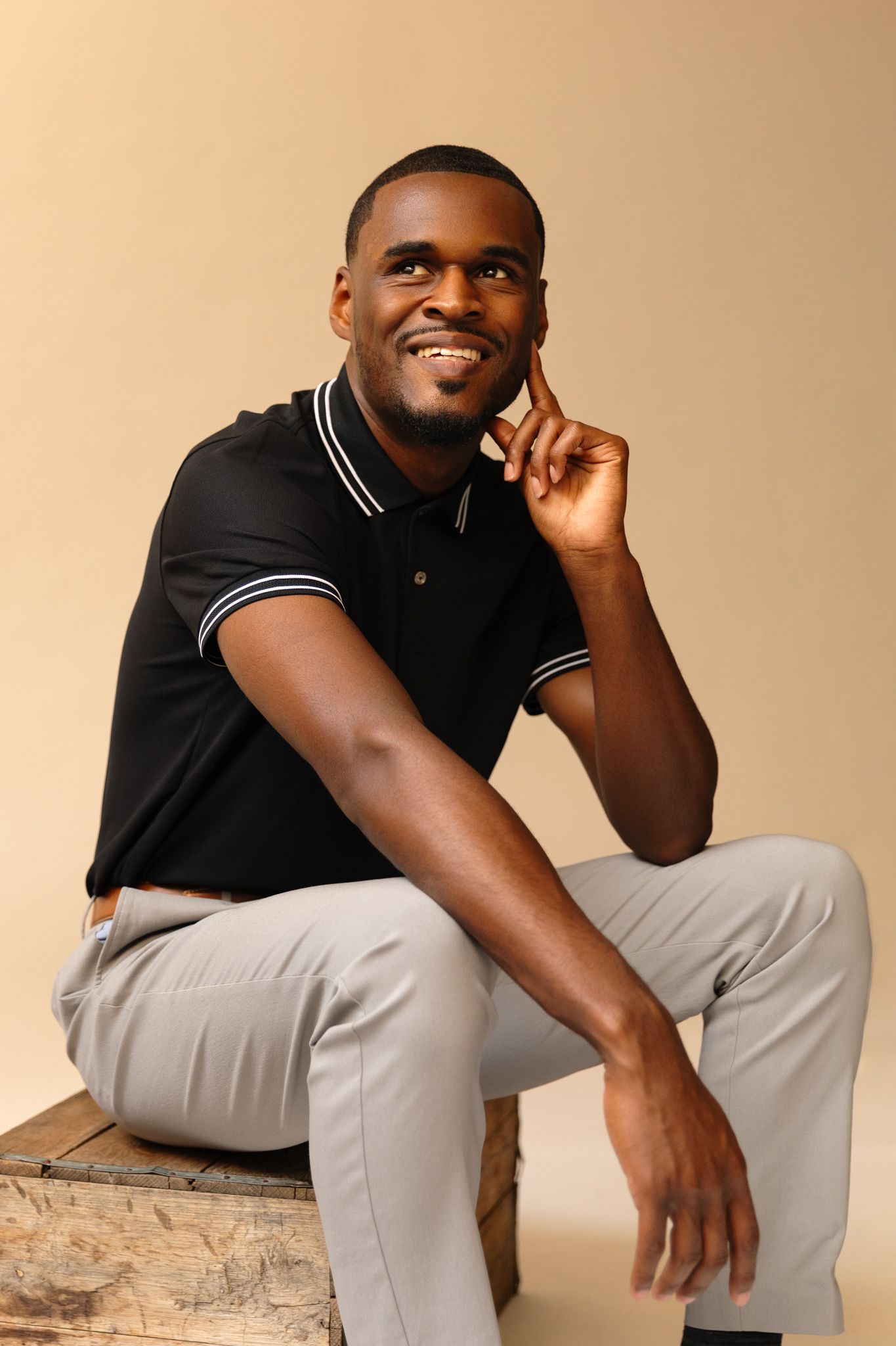 Smiling Black man in black polo shirt sitting on wooden crate against beige background.