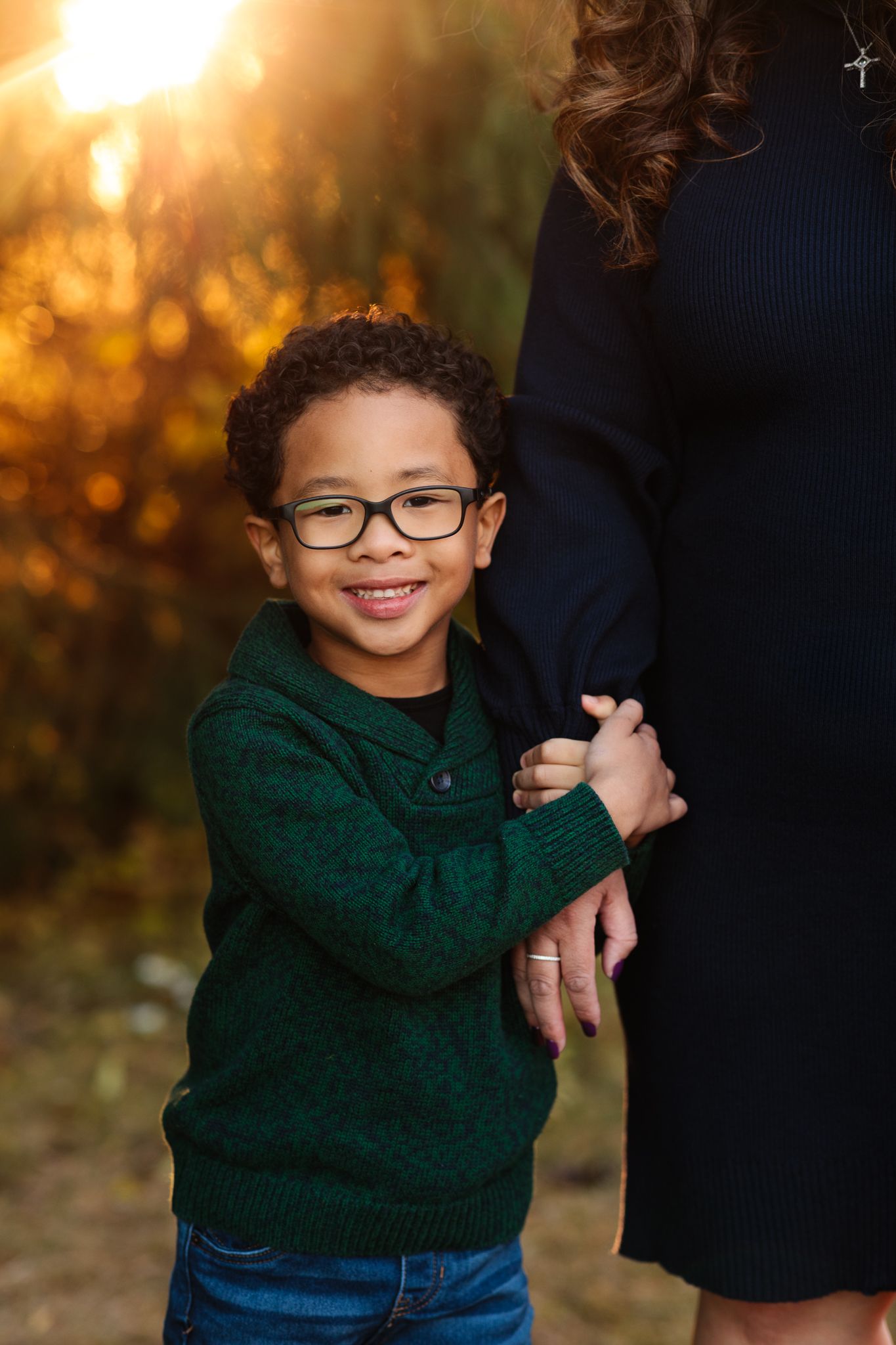 Smiling young boy with glasses in a green sweater hugs his mothers arm outdoors in golden autumn light.