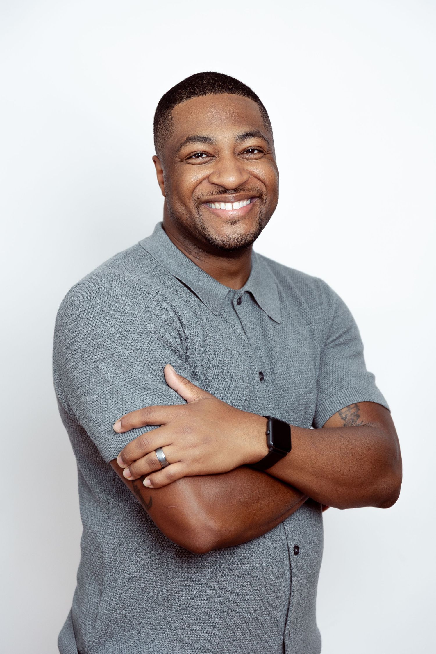 Smiling Black man in gray polo shirt with arms crossed, wearing a smartwatch and ring, posed against white background.