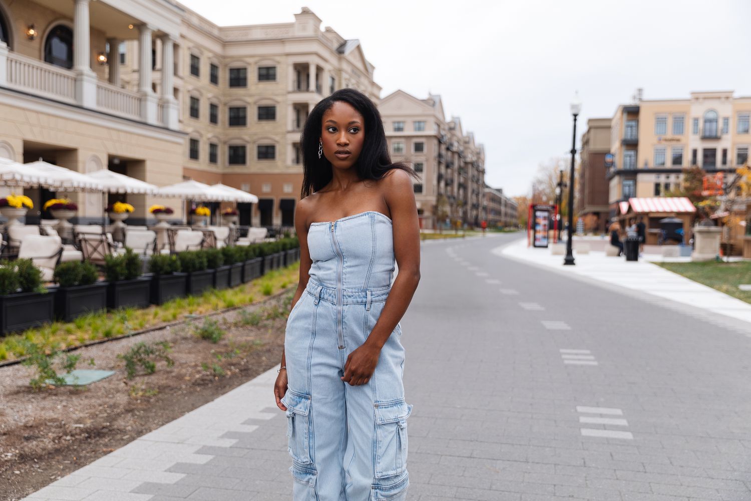 Young woman in denim cargo jumpsuit standing on urban street with buildings behind.