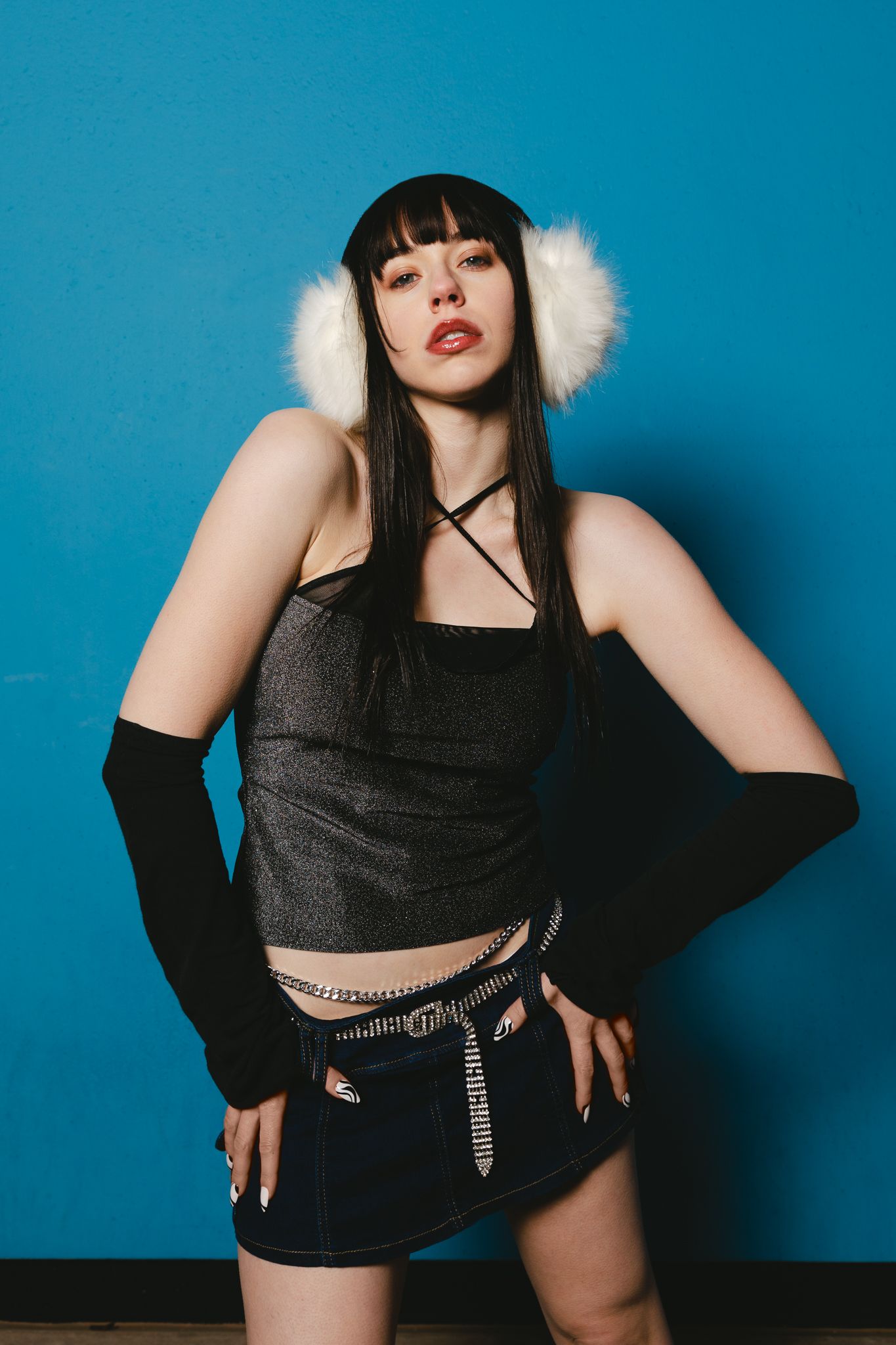 Woman in grey crop top and denim mini skirt wearing white fur earmuffs on blue background.