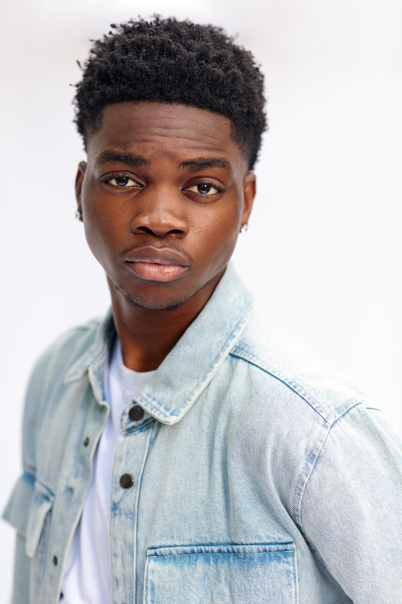 Young Black man in light blue denim jacket posing seriously against white background.