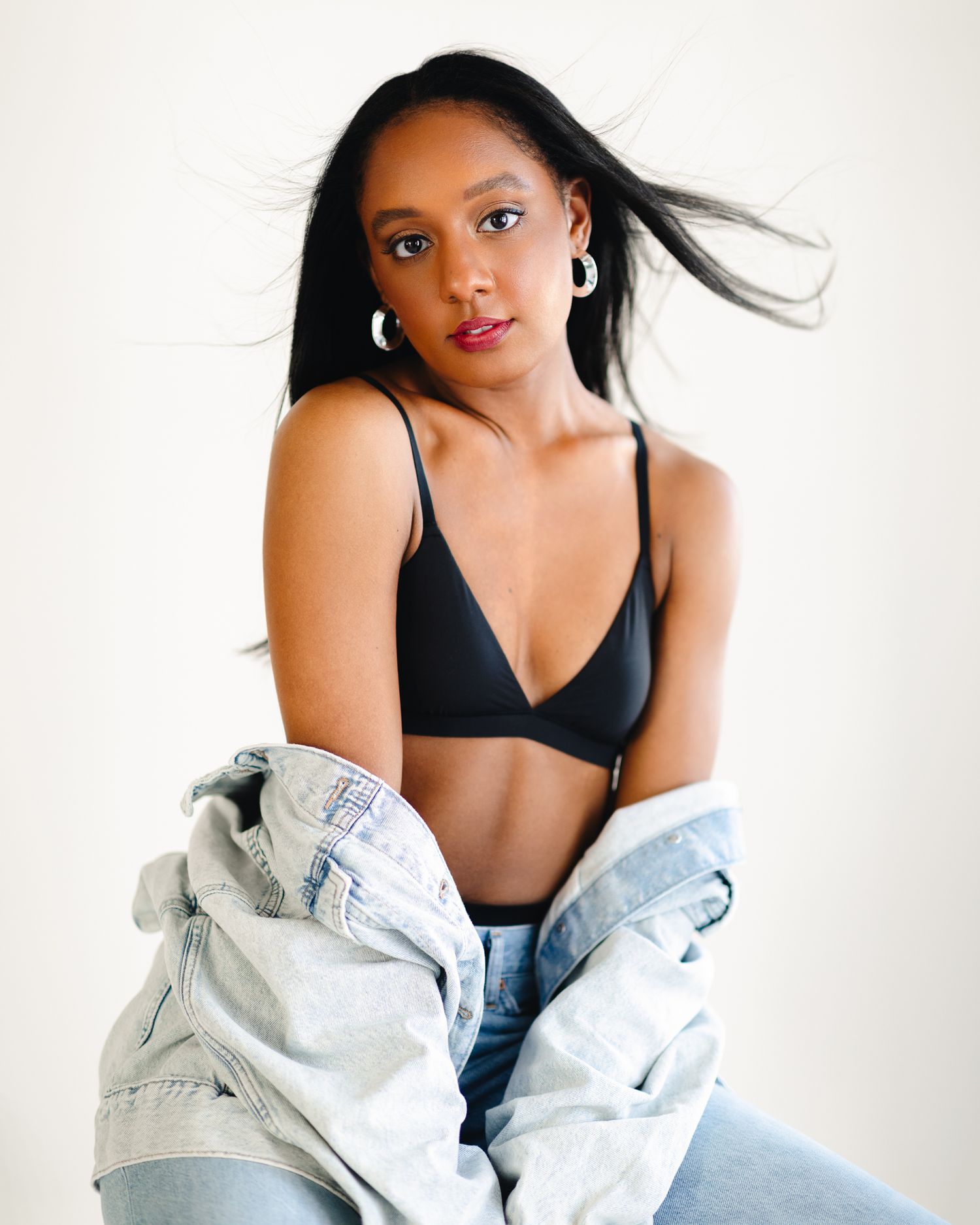 Young woman in black bralette and jeans with denim jacket posing against white background.