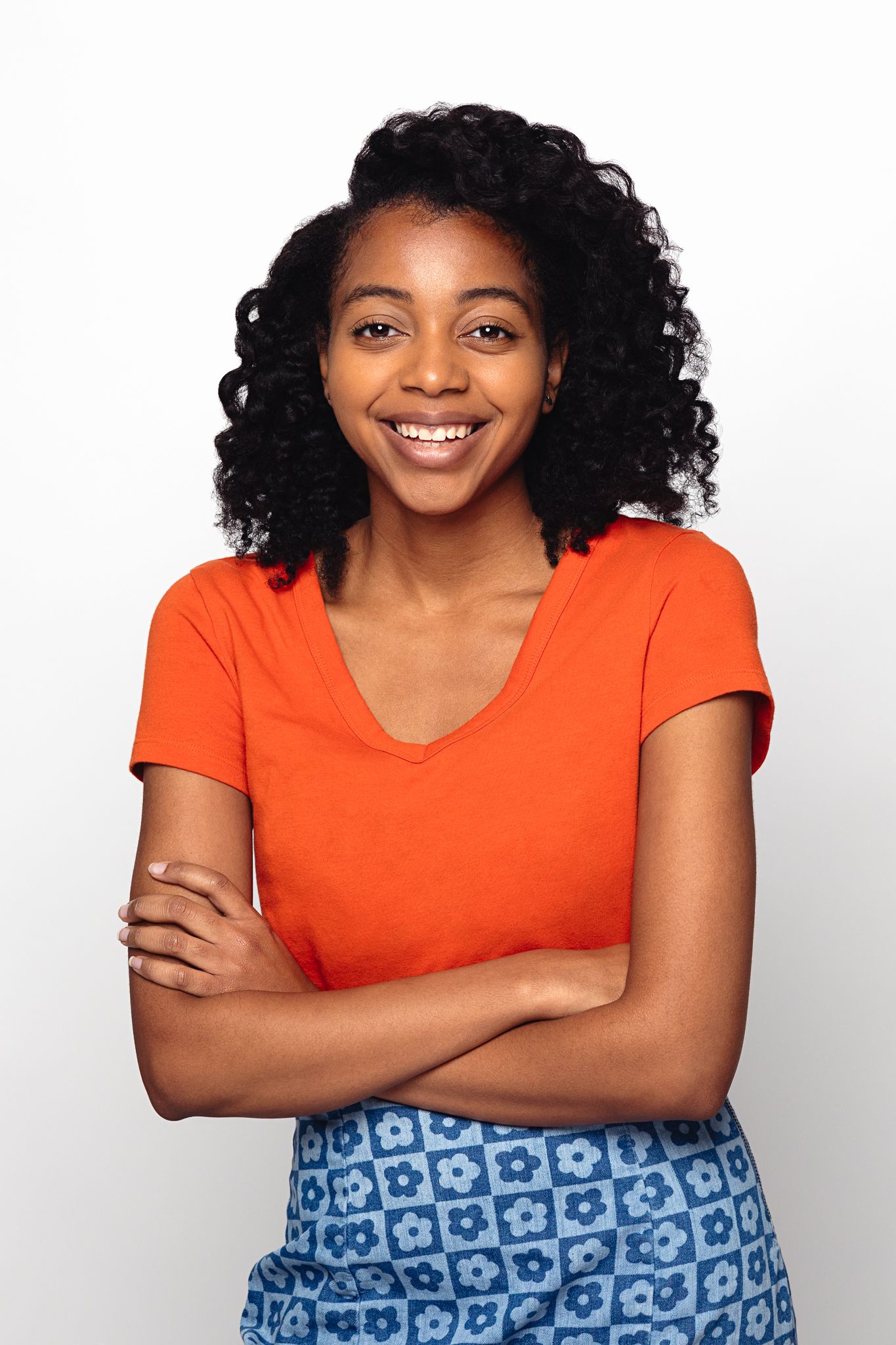 Smiling young woman in orange t-shirt and blue patterned skirt with arms crossed on white background.
