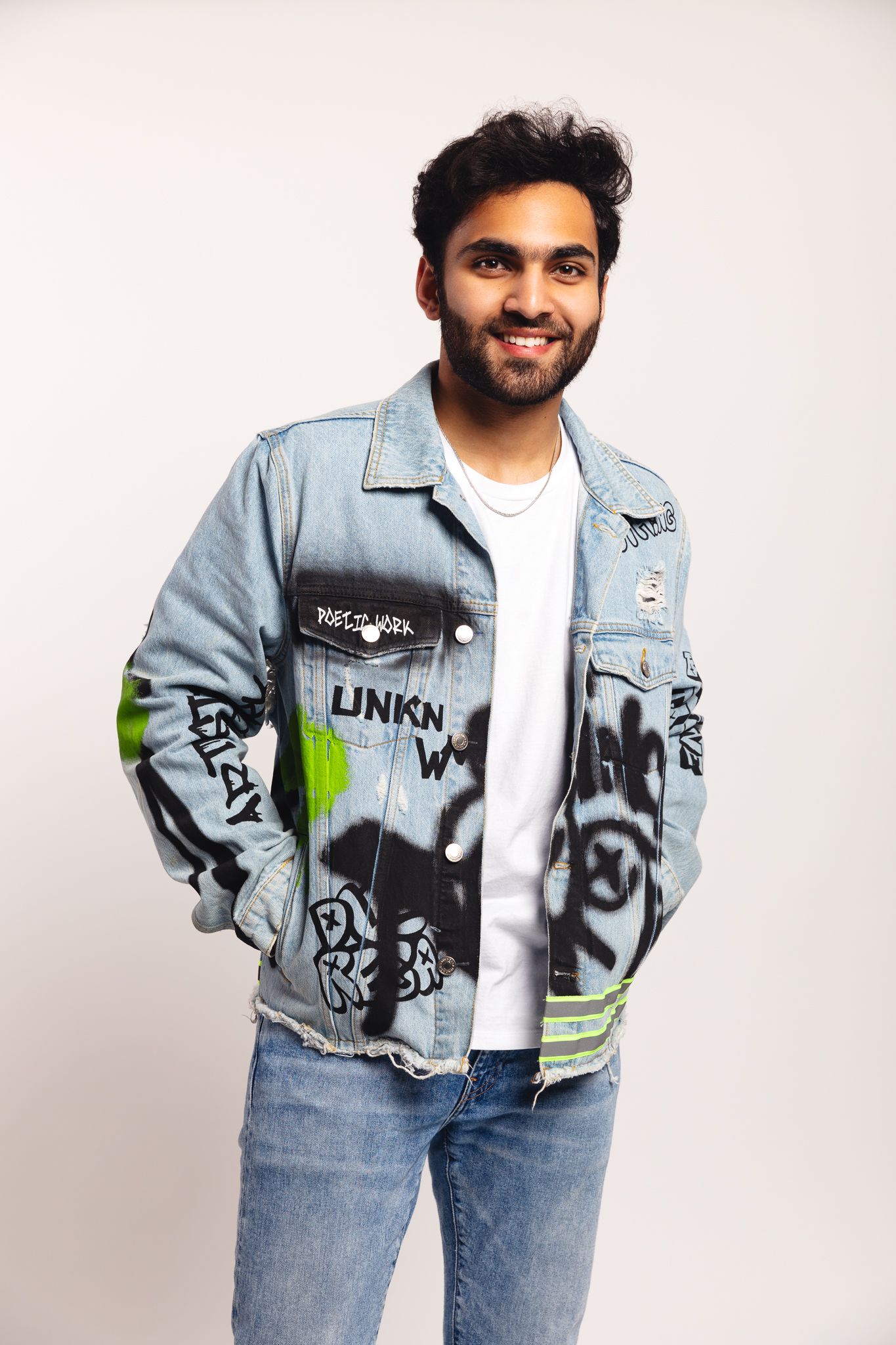 Smiling young man in graffiti-print denim jacket and jeans posing against white background.