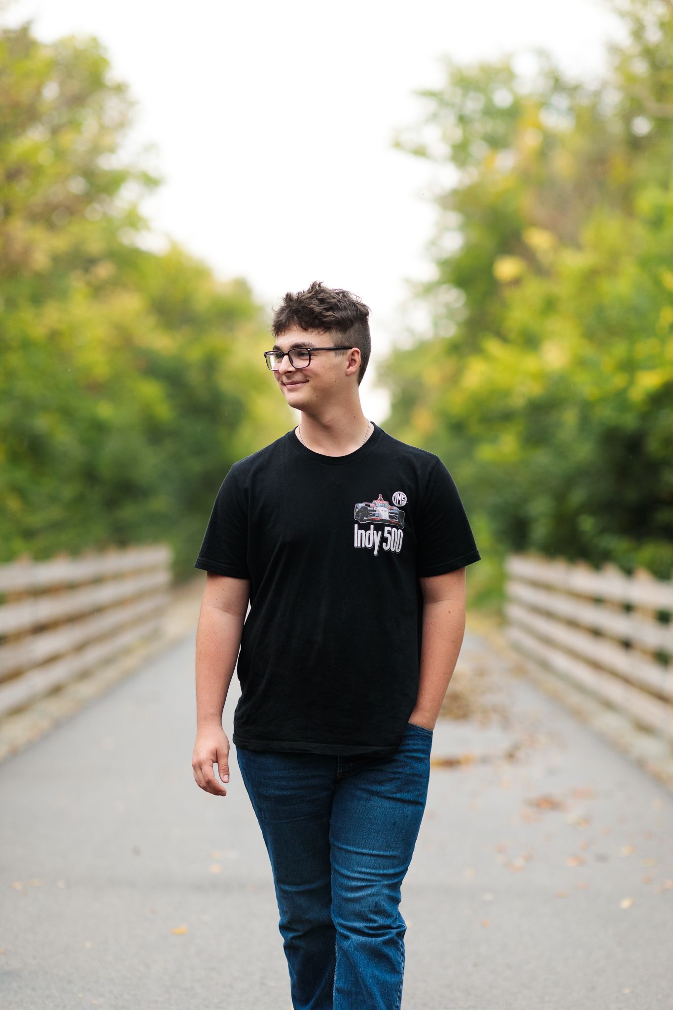 Teen boy in black Indy 500 t-shirt walking on wooden bridge path surrounded by green trees.