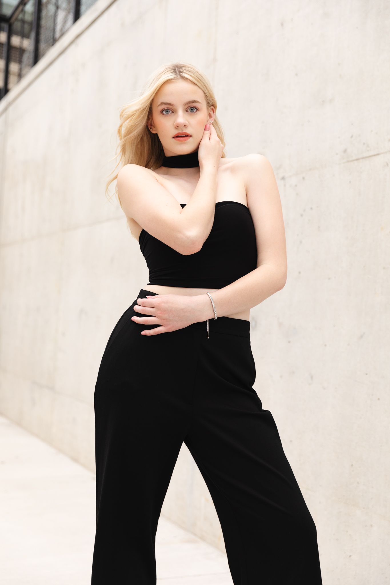Blonde woman posing in black strapless crop top and wide-leg pants with choker necklace against concrete wall.