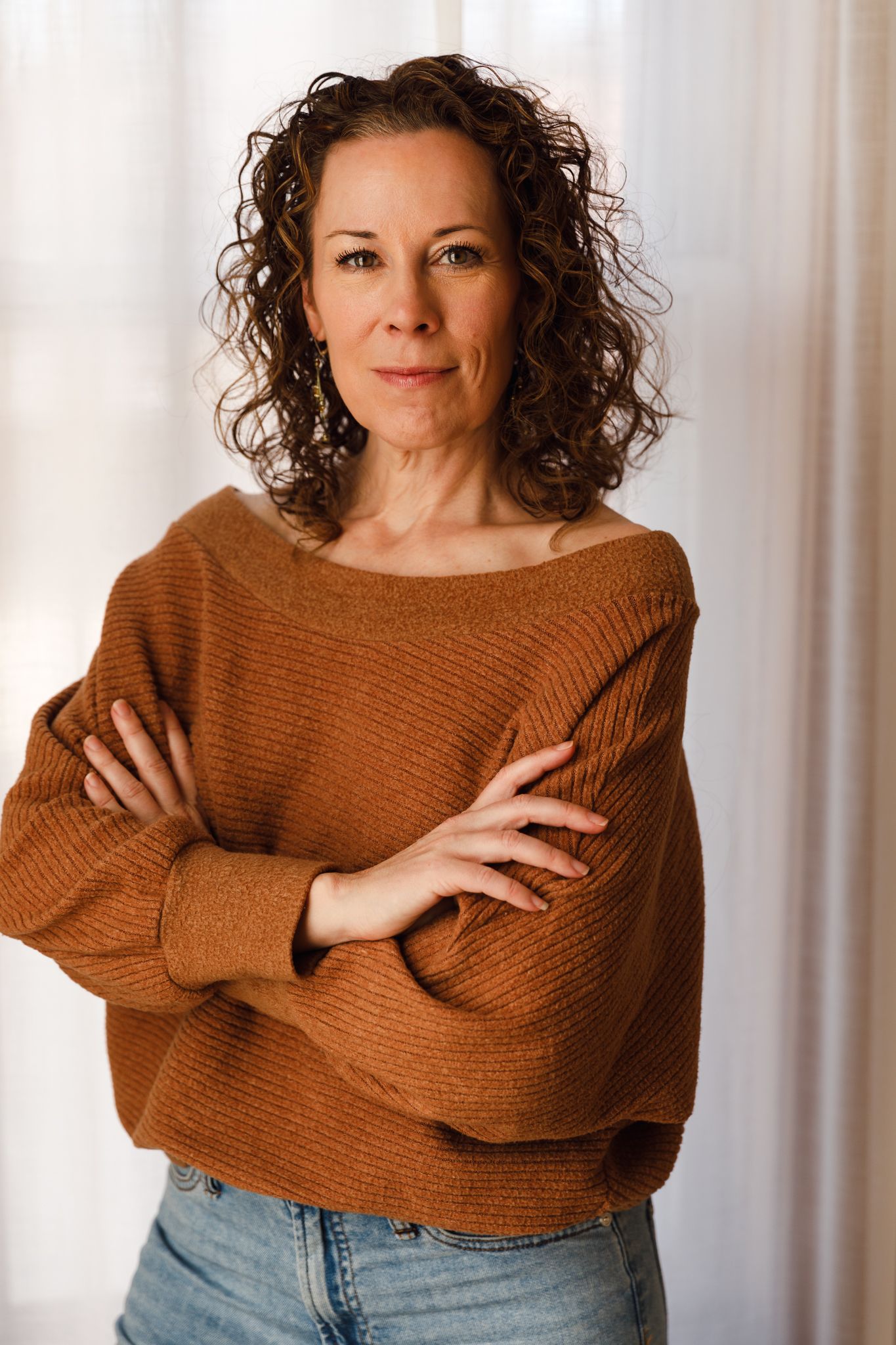 Woman with curly brown hair wearing a rust-colored off-shoulder sweater, standing with arms crossed before a white curtain.