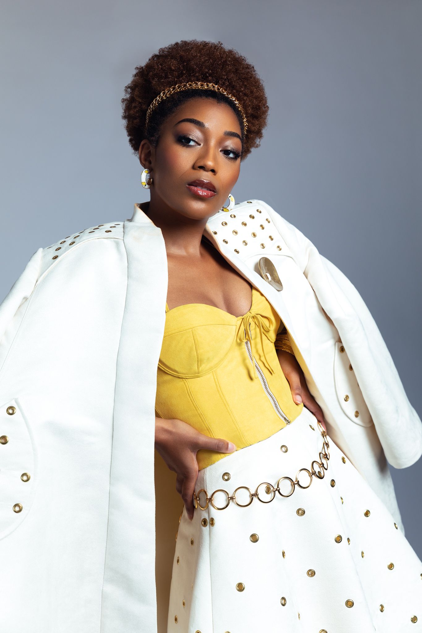Woman in yellow bustier and white studded jacket with gold chain headband in studio.