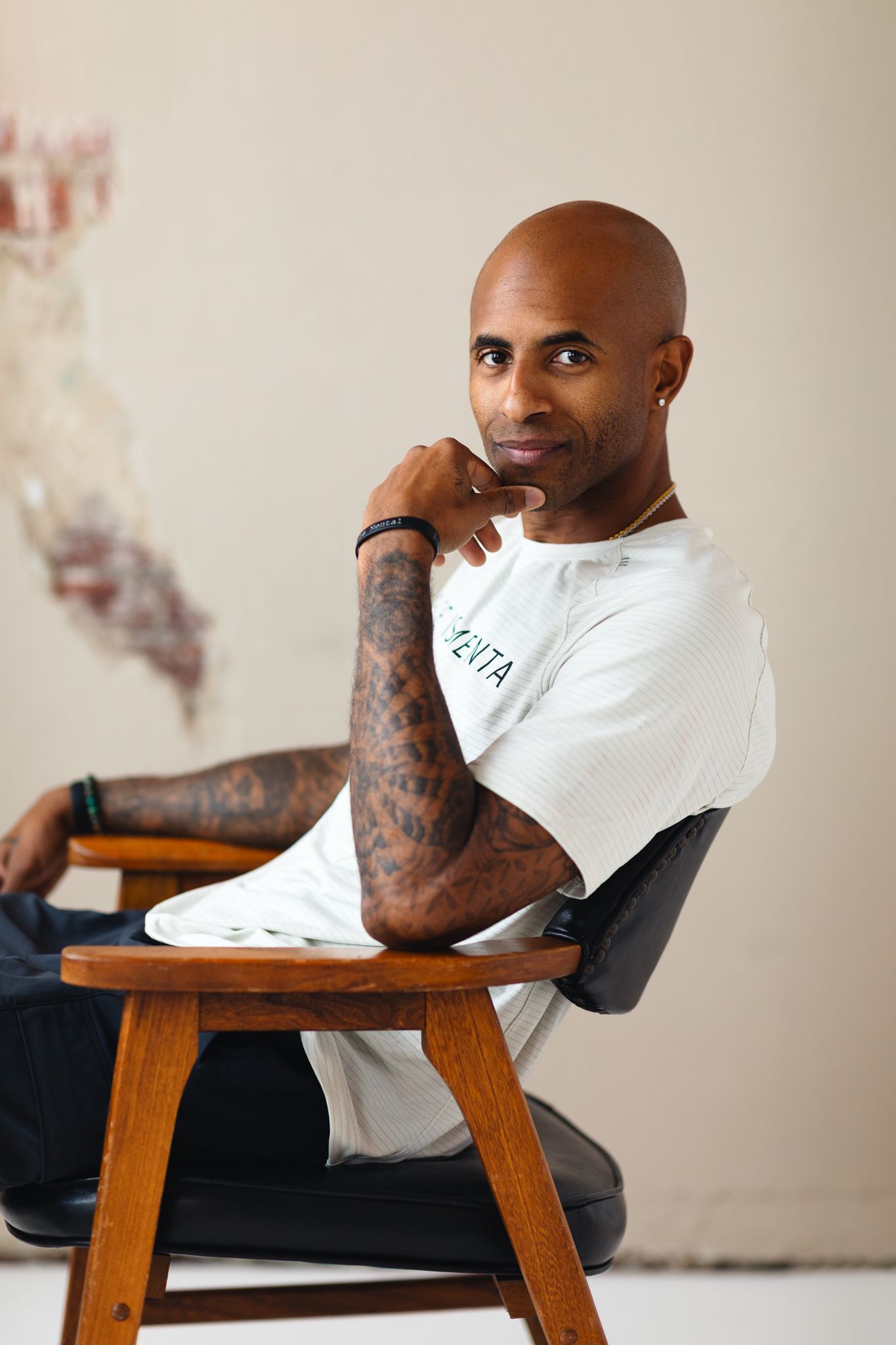 Bald man with tattoos sitting on wooden chair in white t-shirt, posing thoughtfully with hand on chin.