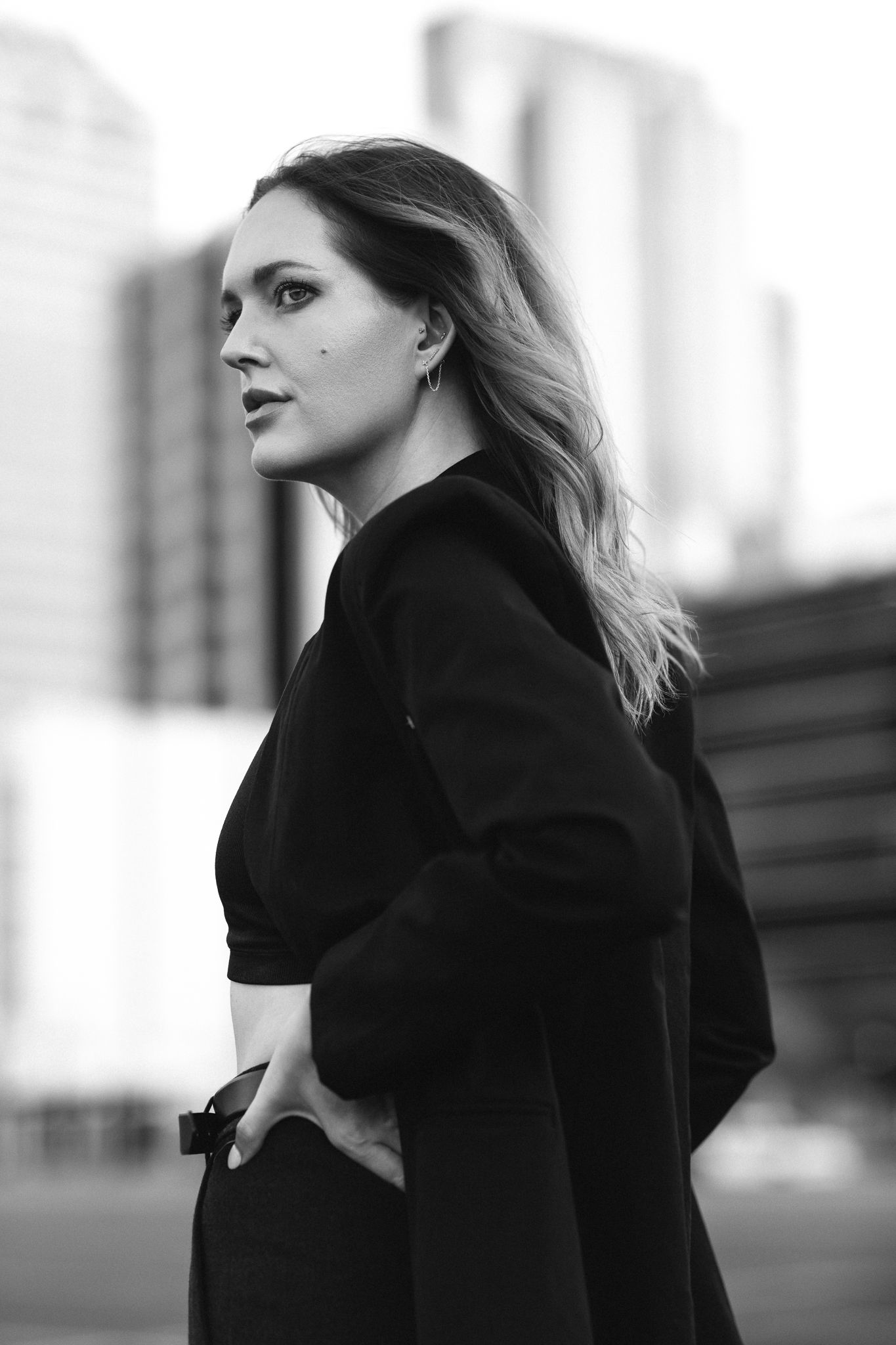 Blonde woman in black outfit posing outdoors with city skyscrapers in background.