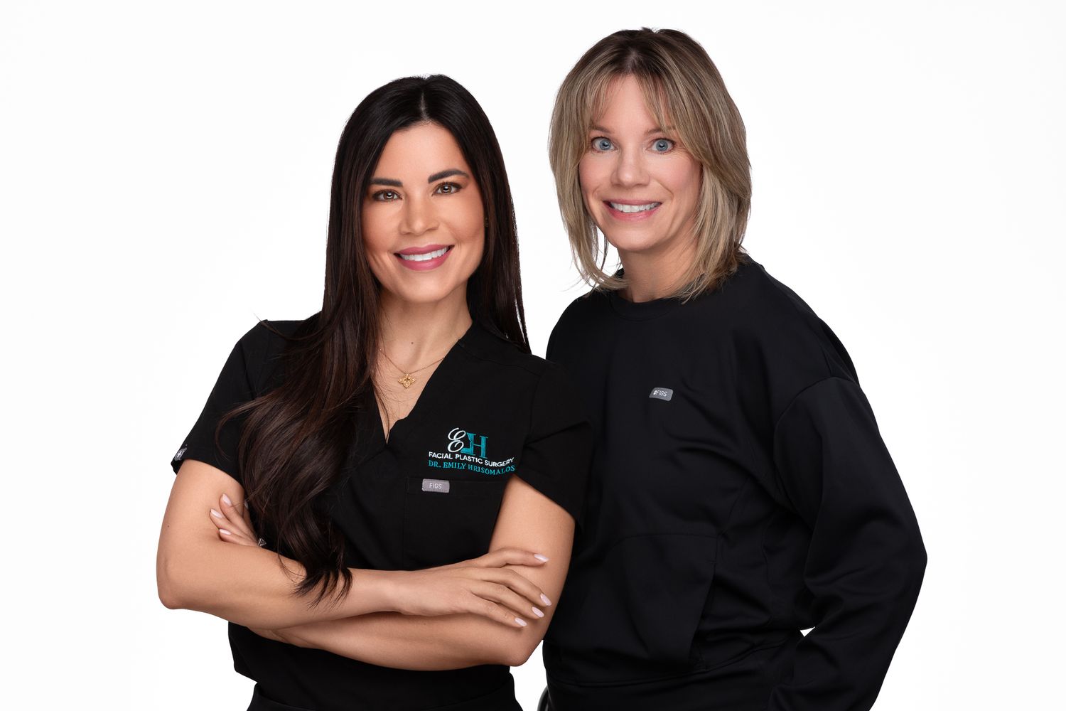 Two women in black medical scrubs pose confidently against a white background in a professional healthcare portrait.