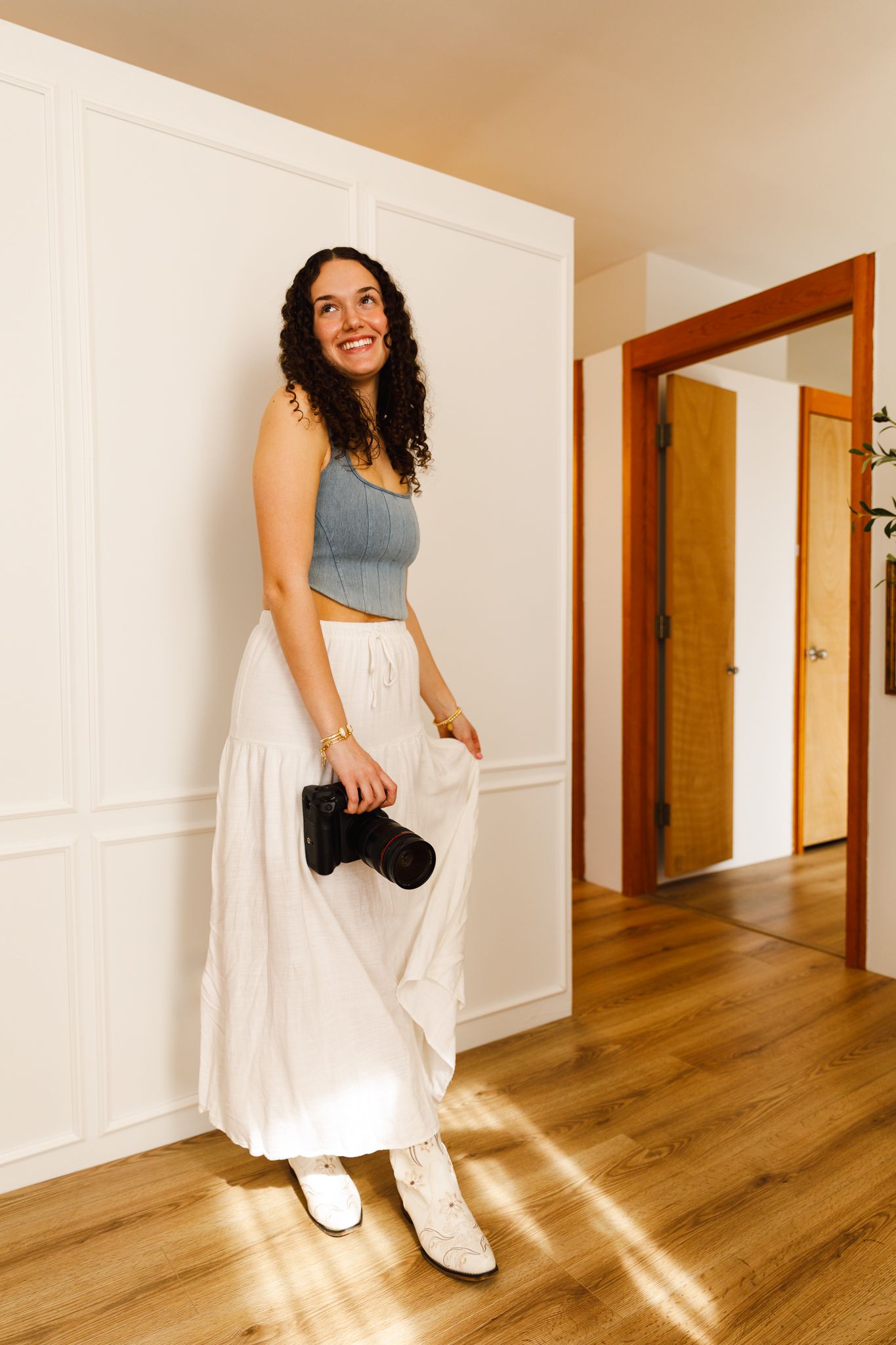 Young woman with curly hair holding a camera, wearing a gray crop top and white maxi skirt, standing in a bright hallway.