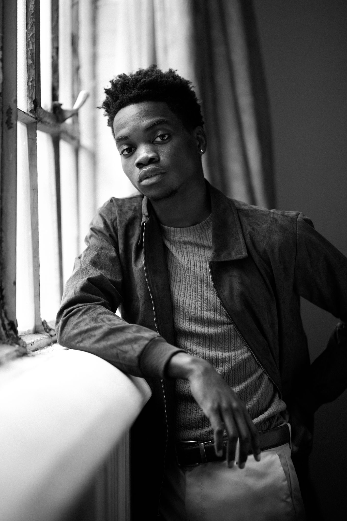 Stylish young man in leather jacket poses by window in black and white photo.