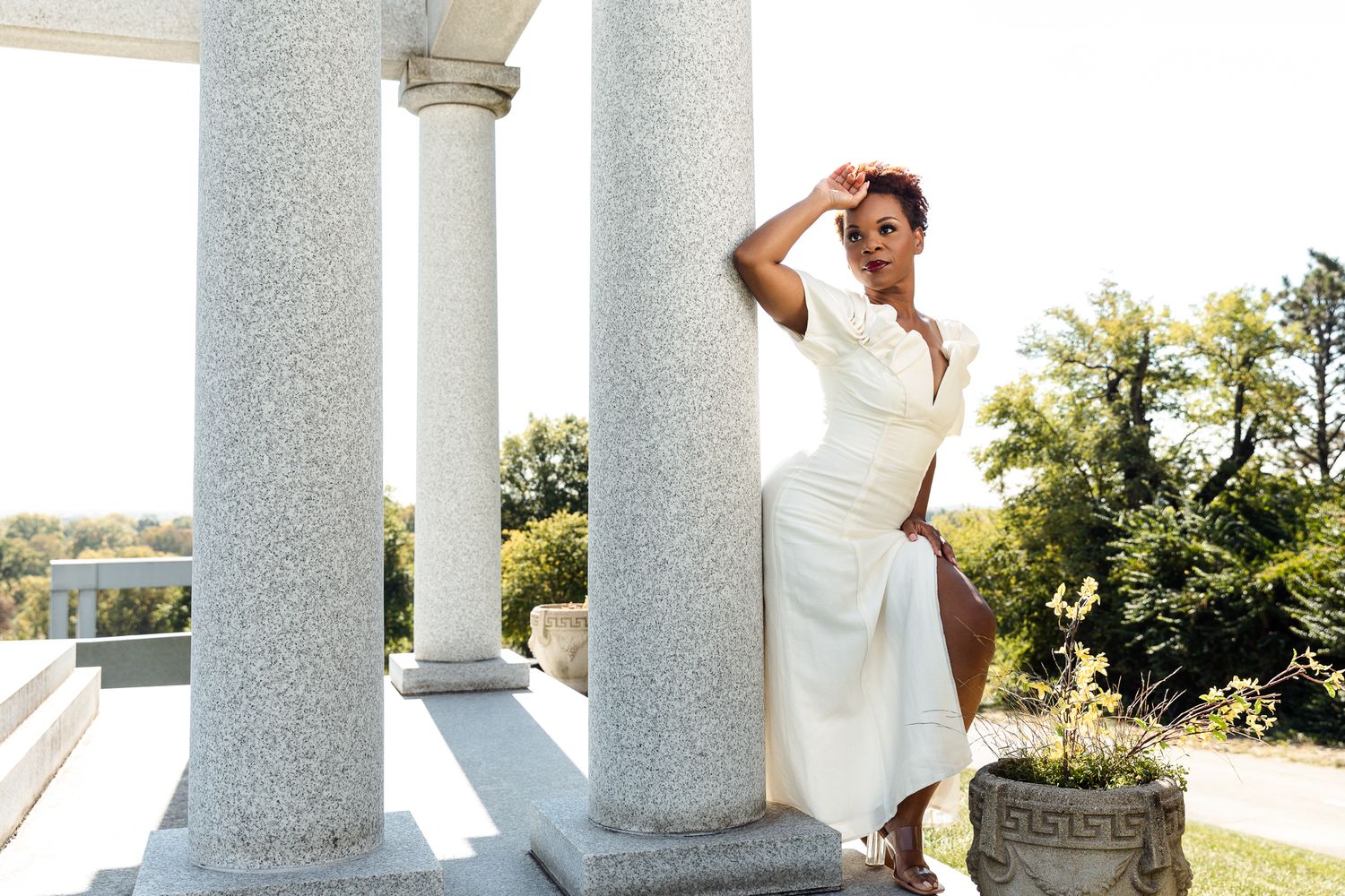 Elegant woman in white dress posing between marble columns at a classical outdoor venue.