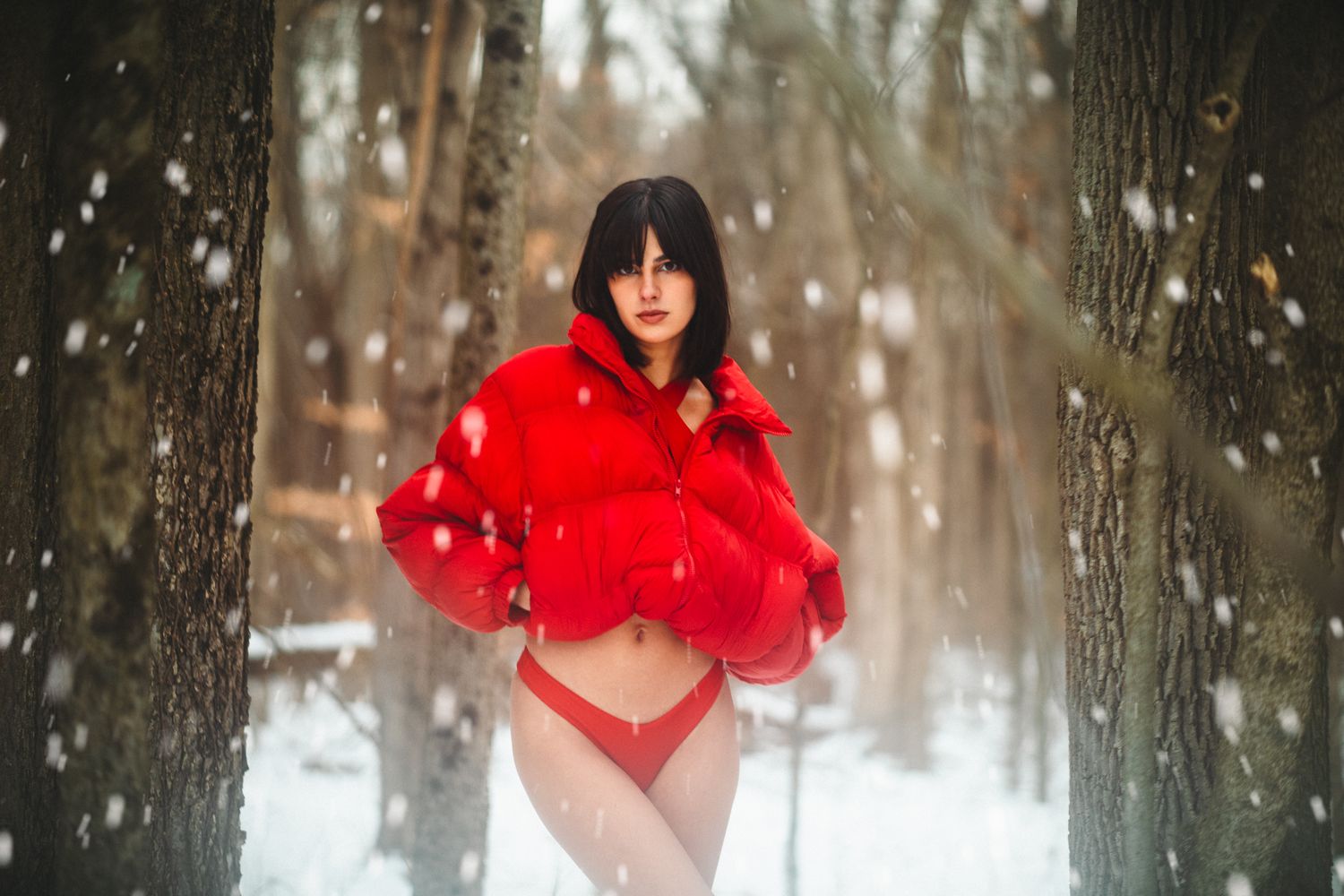 Woman in red puffer jacket and red bikini bottom standing in snowy forest.