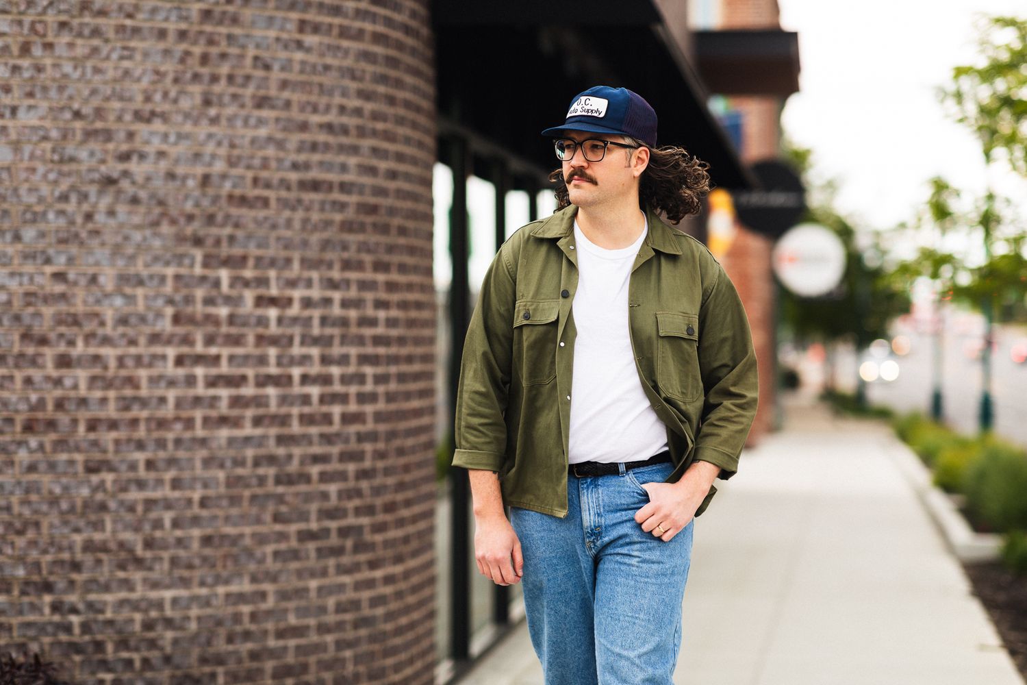 Man in green jacket, white tee, jeans, and navy cap stands on urban sidewalk near brick column.