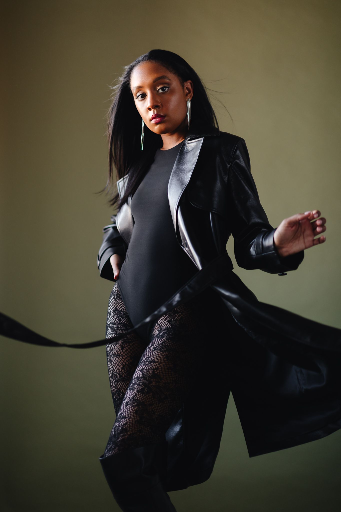 Woman in black leather coat and lace pants posing dynamically on dark background.