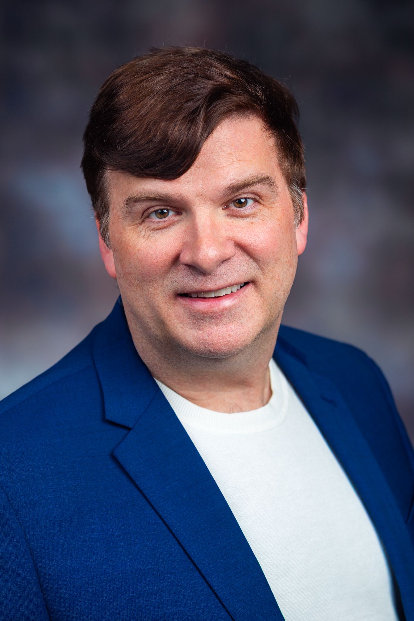 Professional headshot of a smiling middle-aged man in a blue blazer and white shirt against a blurred background.