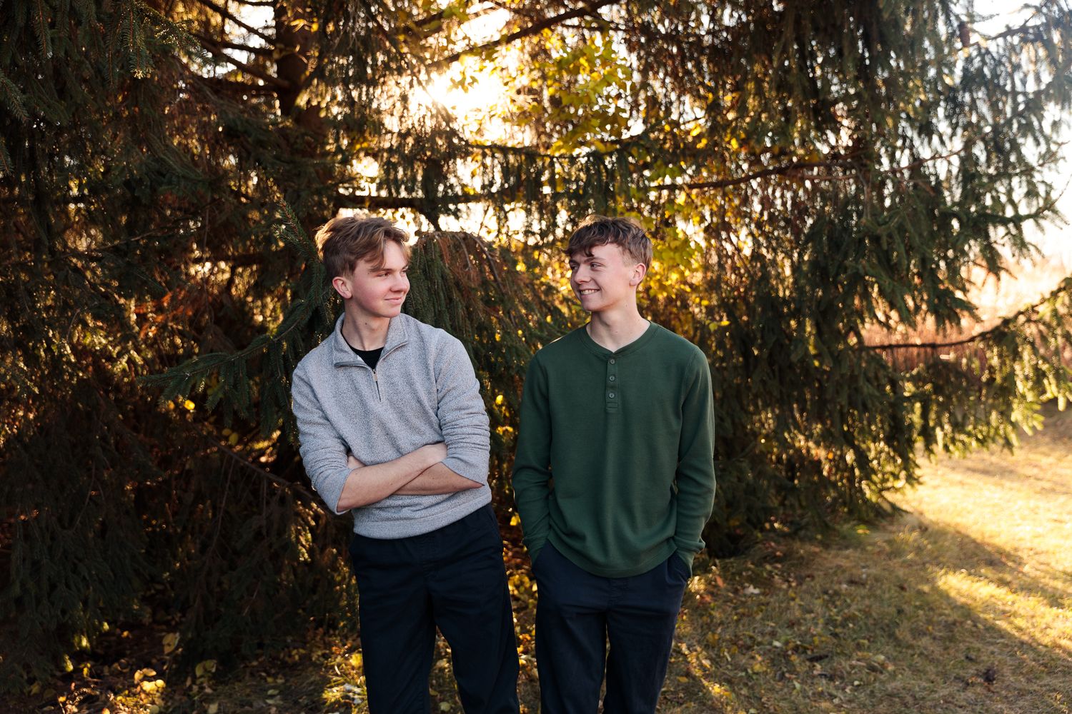 Two teenage boys standing near evergreen trees in golden afternoon sunlight outdoor portrait.