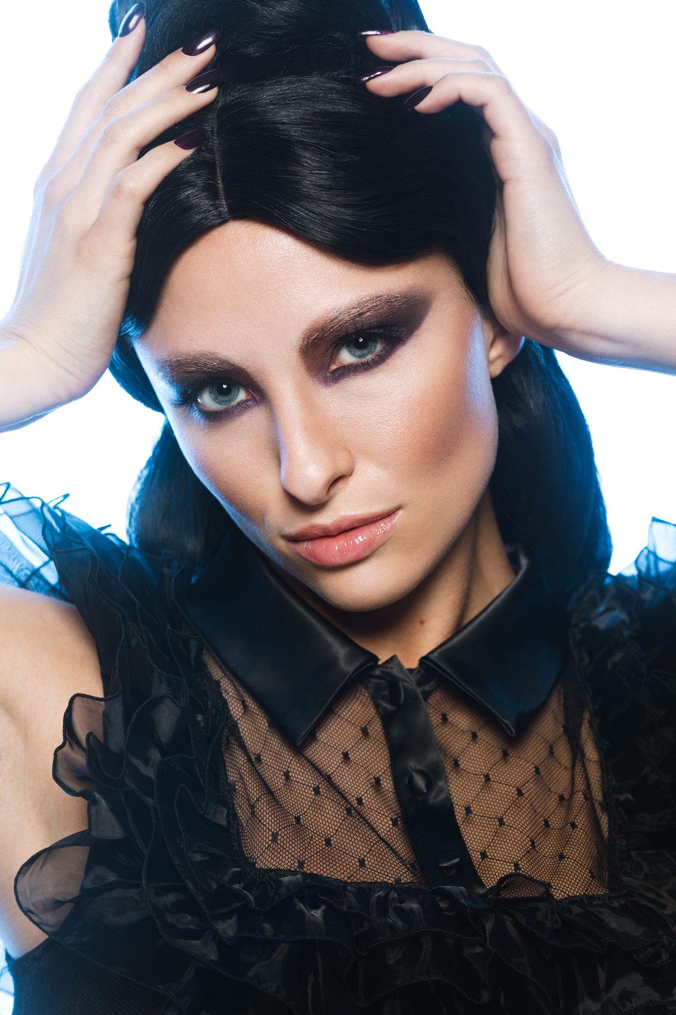 Woman with dramatic dark eye makeup in black ruffled outfit on white background.