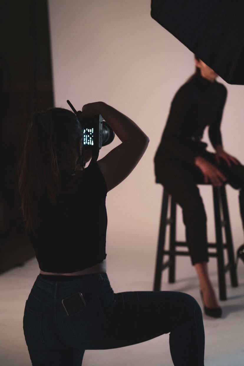 Photographer crouching and shooting a model seated on a stool in a professional photo studio.