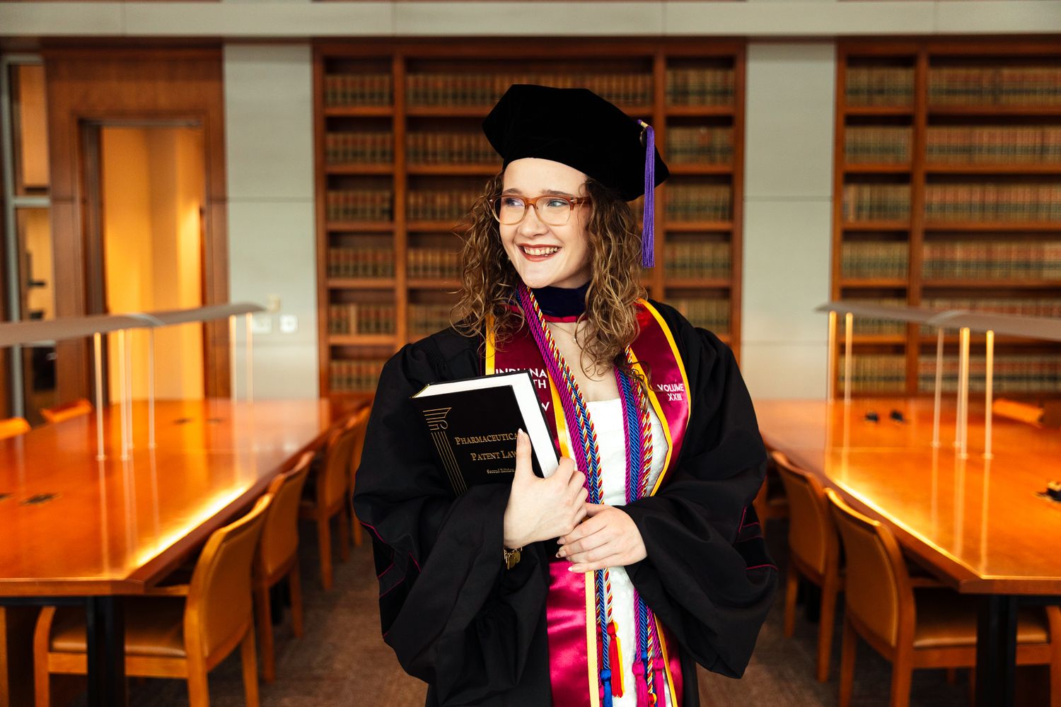 Woman in graduation regalia holding books in a law school library, wearing cap and gown with colorful hood.
