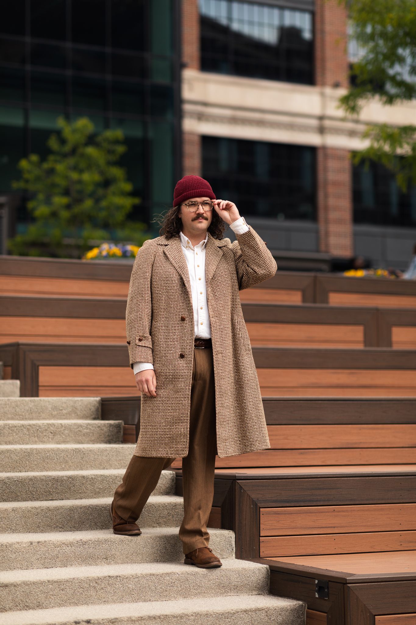 Man in tweed coat, burgundy beanie, and glasses stands on outdoor steps near a modern building.