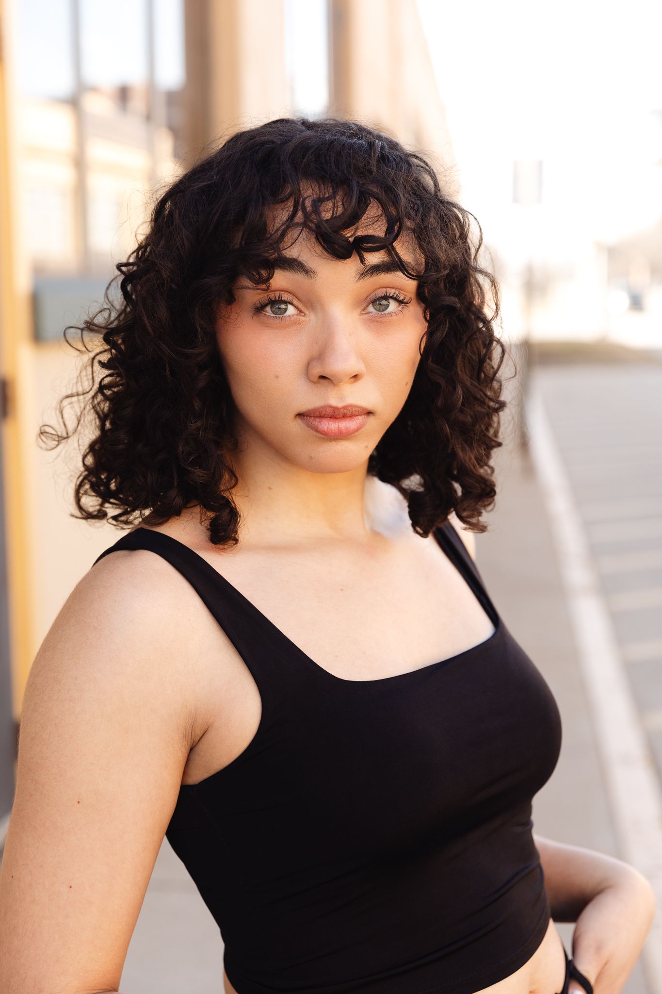 Young woman with curly black hair wearing a black tank top poses outdoors in urban setting with warm sunlight.