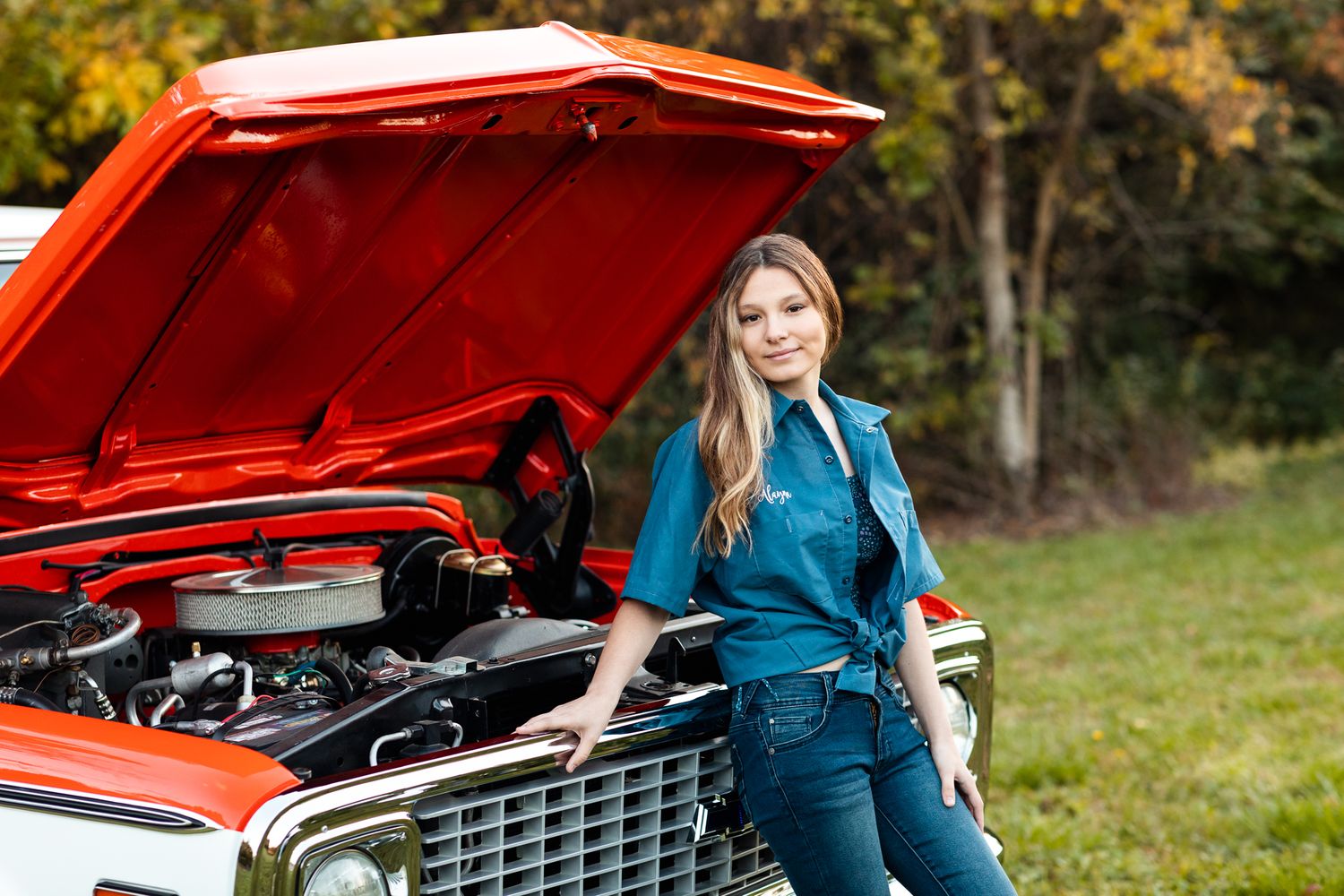 Young woman in teal jumpsuit posing beside classic red car with open hood in outdoor setting.